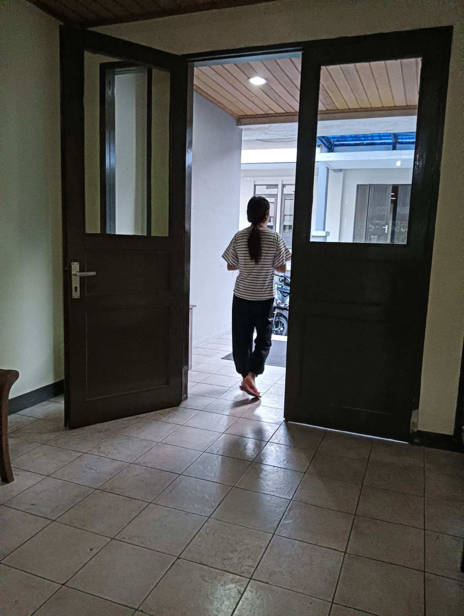 An individual with dark hair pulled into a ponytail, wearing a striped t-shirt and dark pants, walks away from the camera through an open doorway. The person is barefoot and appears to be entering a courtyard area where a motorcycle is parked. The doorway has dark wooden frames and two doors, one of which is partially open. The interior space is tiled with large, light-colored square tiles. The exterior area visible through the doorway has a white building with a covered outdoor space and a blue translucent roof. The ceiling inside the doorway is paneled with light-colored wood. The overall scene suggests a residential setting in Tomohon, Indonesia, likely a home with a courtyard.