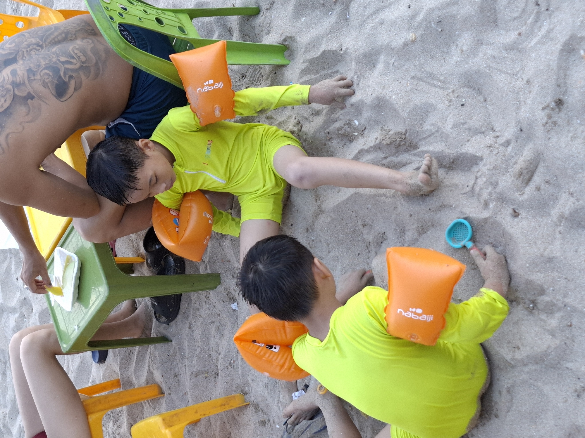 The image shows a high-angle view of two young children in neon yellow rash guards and orange arm floaties lying on a sandy beach. The children are facing away from the camera, with their heads turned slightly. The child on the left appears to be sleeping, while the child on the right is reaching for a blue plastic toy scoop.
In the background, a person with a large tattoo on their back is partially visible, sitting in a low green plastic chair. A yellow plastic tray with a white container of food and a utensil is also on the chair. Other yellow plastic chairs are visible on the sand.
The scene is set on a beach, likely during the daytime, given the bright lighting. The sand appears damp in places, suggesting recent water activity. The overall mood seems relaxed and casual, typical of a beach outing.
Visible text on the orange arm floaties includes the brand name "nabaiji" in white lettering. The rash guard on the left child has some white text on it, but it is not clearly legible. The location is identified as Bac Nha Trang Ward, Vietnam.