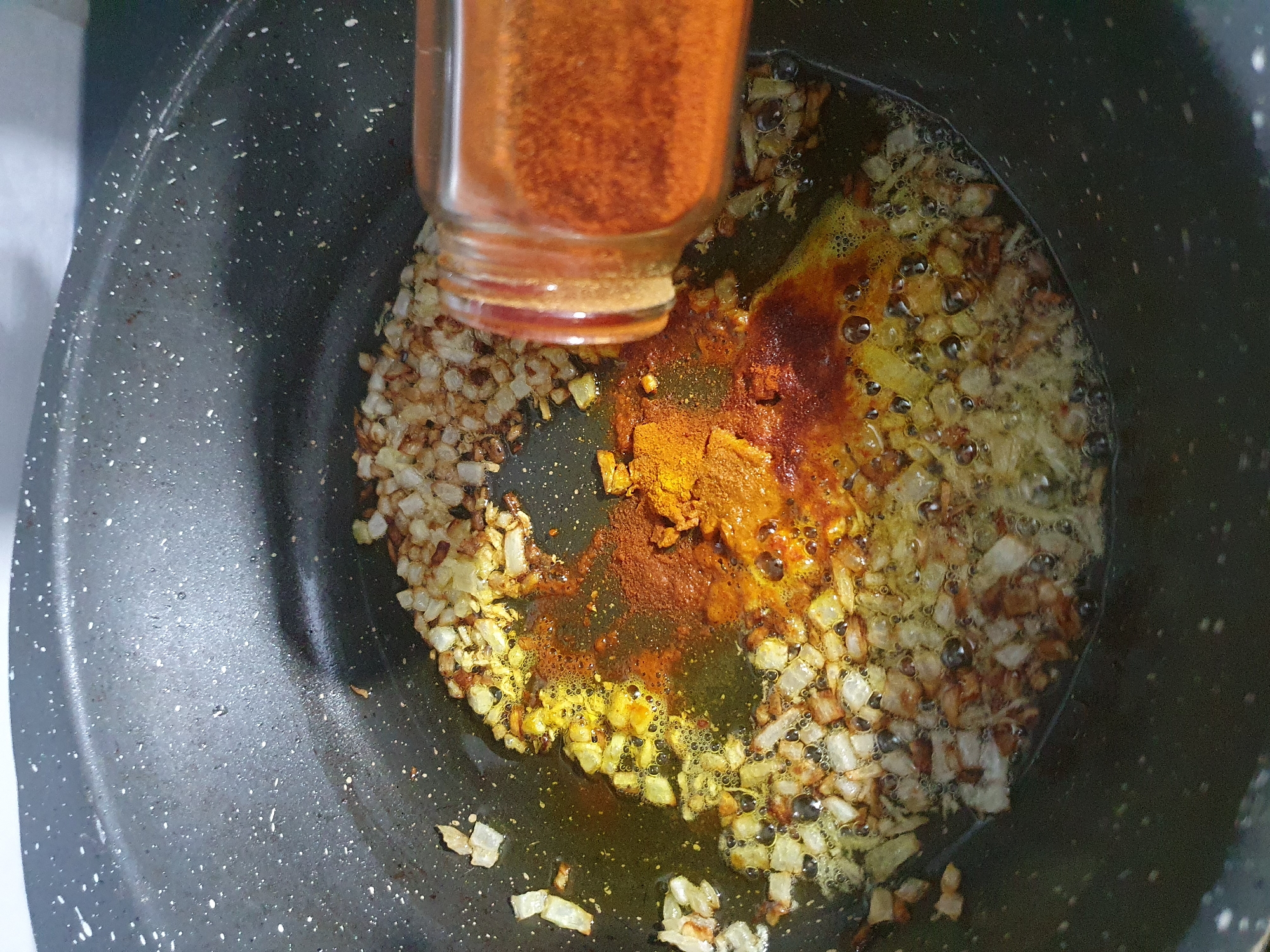 In a dark, non-stick pot, diced onions are sautéed in oil and are beginning to brown. A spice blend, appearing to be turmeric and paprika, is being sprinkled into the pot from a glass jar with an orange-tinted lid. The oil is bubbling around the onions and spices, indicating the heat is on. The background is blurred and out of focus. There is no discernible location in the city of Unknown, Unknown.