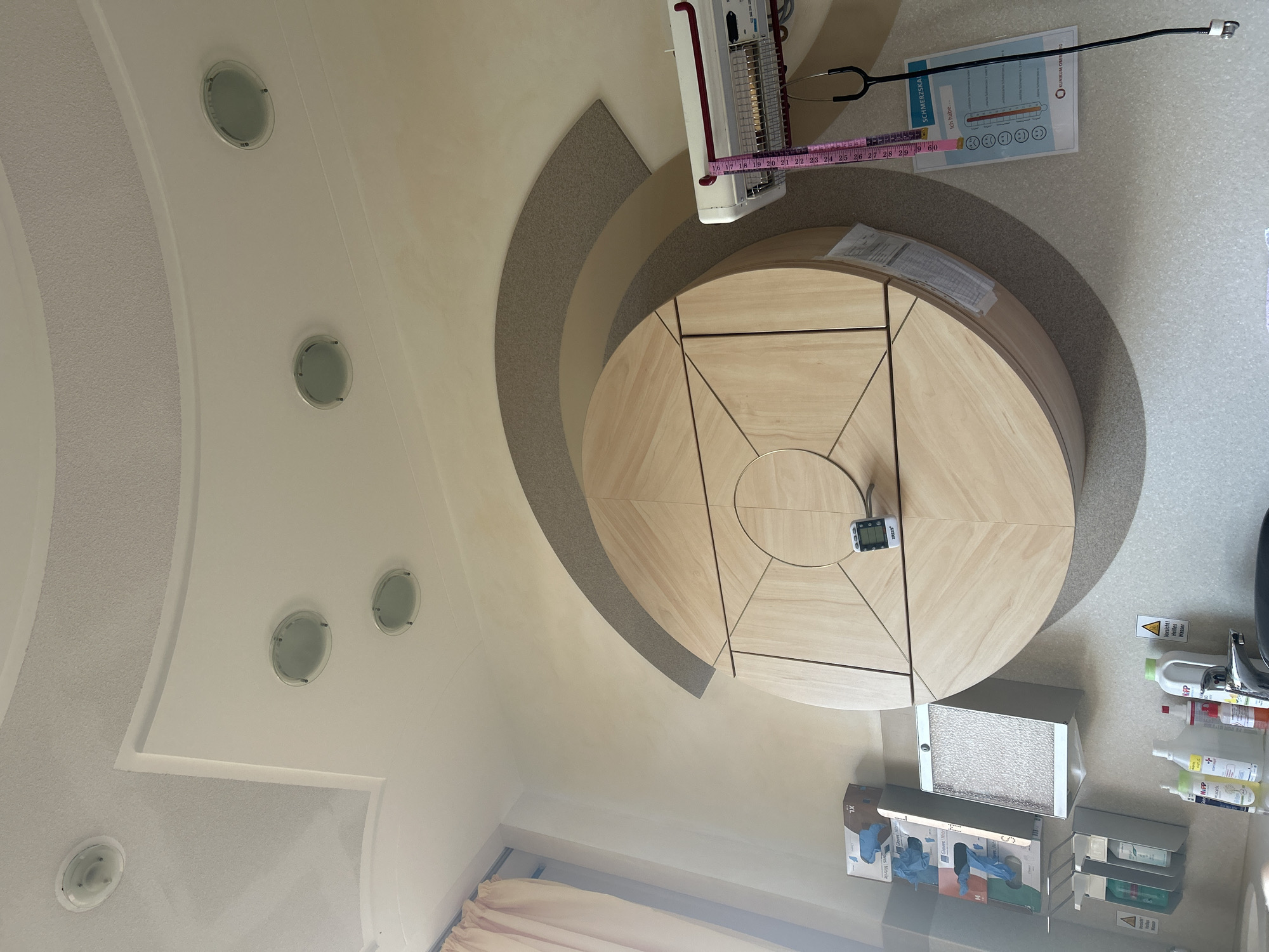 The image depicts an interior scene, likely a medical examination room or office, with a focus on a large, circular wooden cabinet mounted on the wall. The cabinet features a complex geometric design with inlaid wood patterns. Above the cabinet, the ceiling has a multi-level design with recessed circular lighting fixtures. To the left of the cabinet, there are wall-mounted dispensers for gloves and hand sanitizer, as well as boxes of medical gloves. A curtain is visible to the far left. To the right of the cabinet, a pink measuring tape hangs next to a medical device, possibly a heater or light therapy unit, and a stethoscope is attached. Below this, a chart with a pain scale and smiley faces is affixed to the wall. The wall surface has a textured, light-colored finish. Several bottles of cleaning or medical supplies are visible on a counter or sink area in the lower left portion of the image. The lighting is bright, suggesting daytime. The overall impression is one of a clean, organized, and functional medical setting. The location of Gummersbach, Germany is not explicitly indicated by any visual cues in the image.