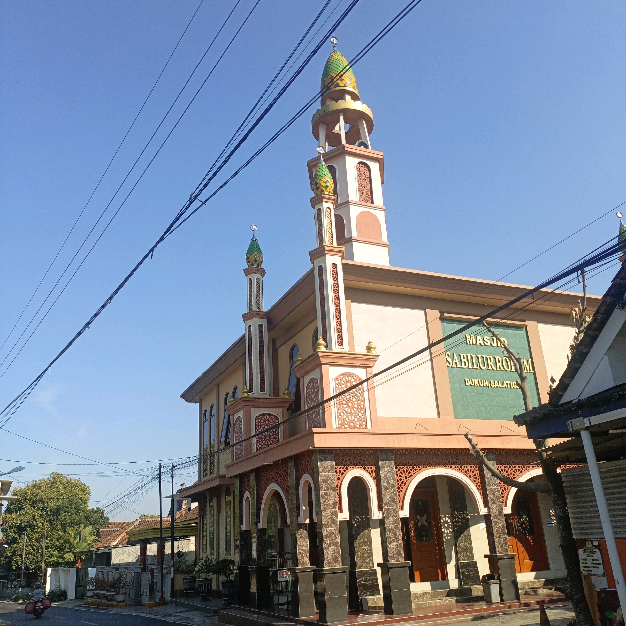 The image shows a mosque with two minarets under a clear blue sky. The mosque has a light brown facade with ornate windows and decorative arches on the lower level. The minarets are tall and slender, topped with green and yellow domes. A sign on the mosque reads "MASJID SABILURROHMAN DUKUH, SALATIGA". The foreground features a street with a person riding a scooter and a few potted plants. Electrical wires crisscross the sky above the mosque. The mosque appears to be situated in a residential or semi-urban area, as suggested by the surrounding houses with tiled roofs visible in the background. The lighting indicates it is daytime, likely morning or afternoon due to the clear sky and shadows.