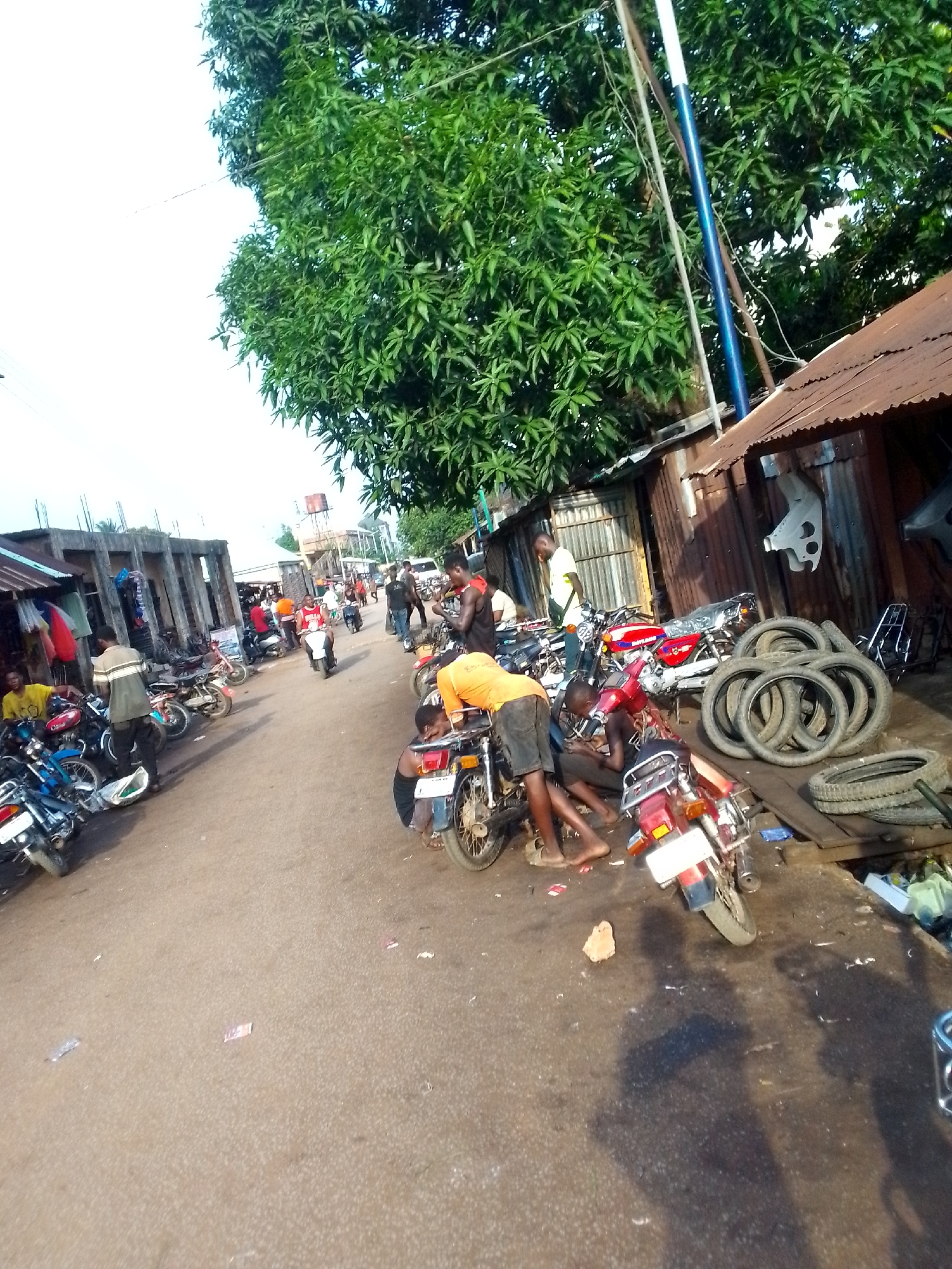 The image depicts a street scene in Orlu, Nigeria. A cluster of men are gathered around several motorcycles that are parked on the side of a paved street. The men appear to be working on the motorcycles. To the left, are several businesses that sell goods and motorcycle parts. A large tree is present in the scene and provides shade for the right half of the scene. A stack of tires is visible on the right. A couple of electrical poles can be seen as well. In the distance, other people are walking down the street.