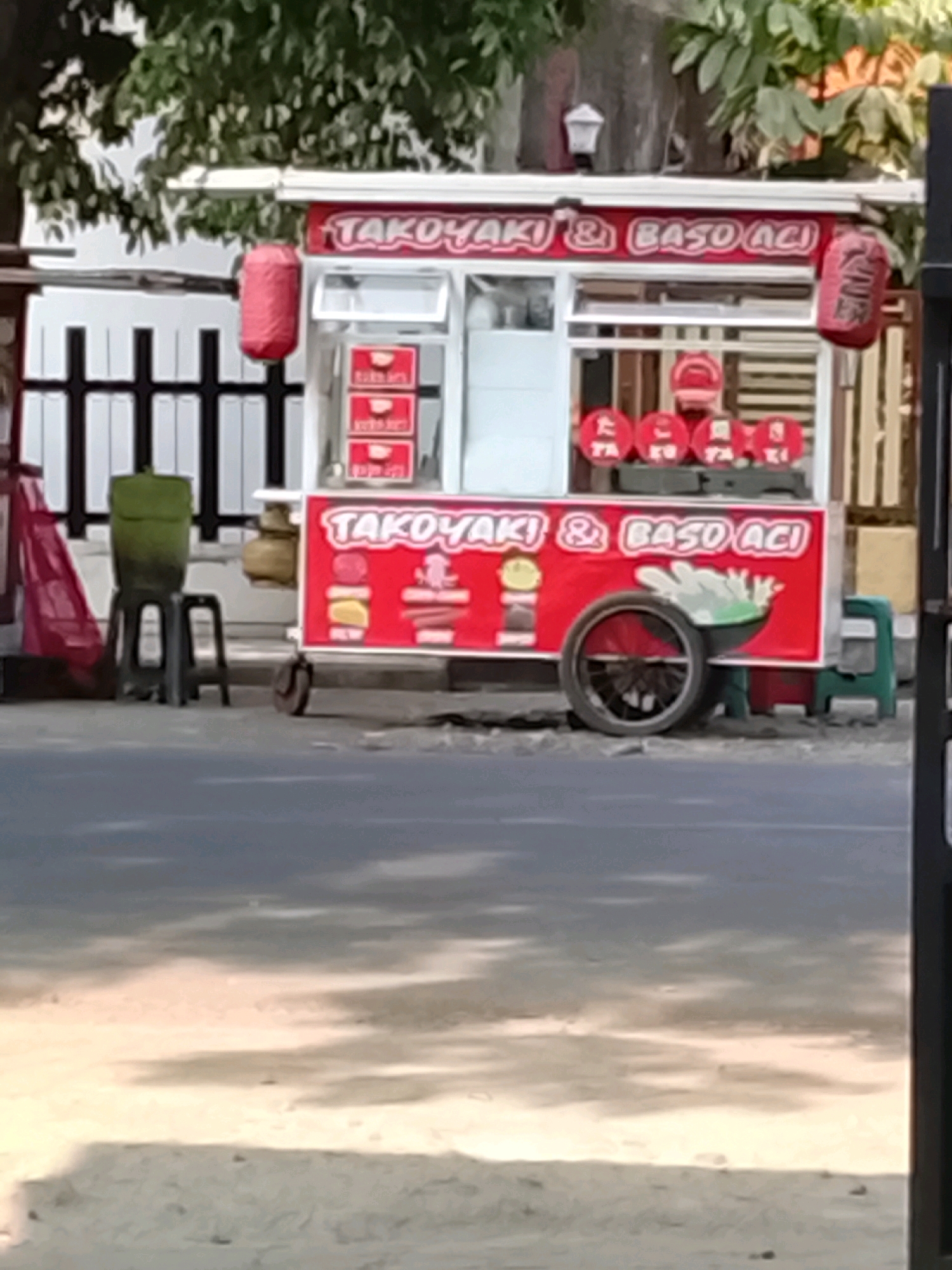 A food cart is parked on the side of a street in Slawi, Indonesia. The cart is red and white and has signs advertising "Takoyaki & Baso Aci." There are red lanterns hanging from the sides of the cart. In front of the cart, there are three red circular signs with Japanese characters. Behind the cart, there is a black fence and some greenery. The ground is paved with asphalt.