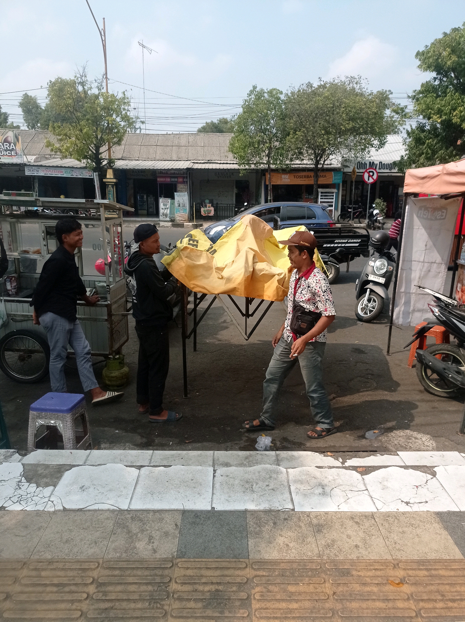 In the city of Tuban, Indonesia, three men are engaged in activity near a food cart. One man, wearing a black shirt and light blue pants, stands by a food cart and smiles. Another man, dressed in a black hoodie and black pants, stands near a covered table with a yellow tarp. The third man, wearing a patterned shirt, brown hat, and gray pants, walks past the table, holding a cigarette and a sling bag. In the background, various shops and a blue car are visible, along with motorcycles parked on the street. The scene depicts a lively street-side market or food stall area.