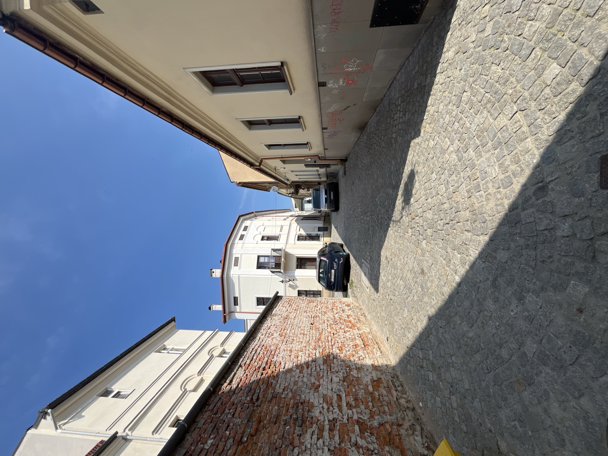 A low-angle, full shot captures a narrow cobblestone alleyway in an urban setting. The alley is flanked by buildings on both sides, with a red brick wall on the left and light-colored buildings on the right. The wall on the left casts a large shadow across the cobblestones. Two cars are parked in the alley: a dark blue car on the left, and a silver van further down on the right. The sky above is a clear, bright blue. The buildings show architectural details such as windows and balconies, and the wall on the right has graffiti. The scene appears to be captured during daytime, with strong sunlight indicating fair weather.