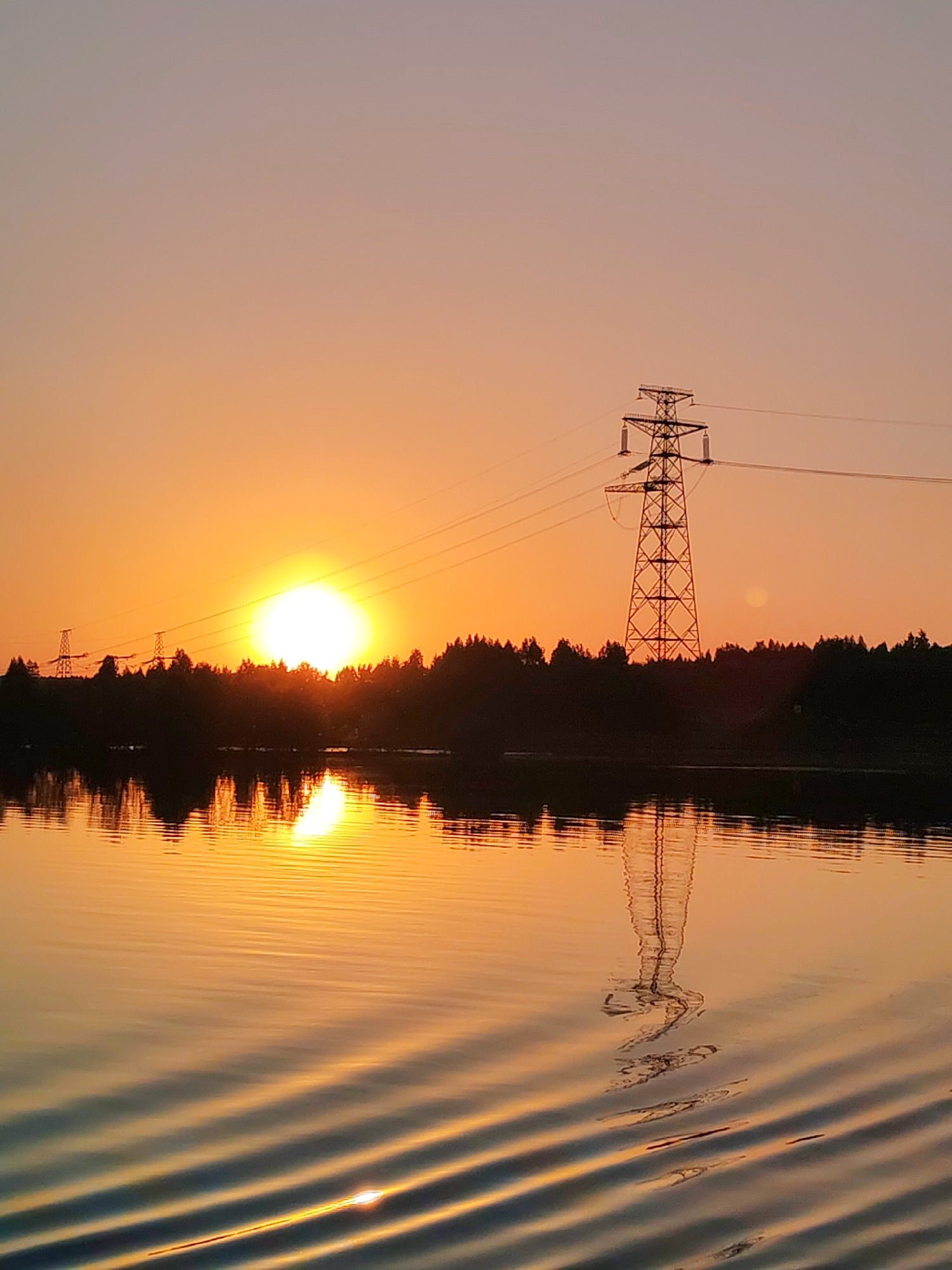 The image shows a sunset over a body of water. The sun is positioned above a line of trees, with its reflection visible in the water. To the right of the sun is a tall transmission tower. Power lines are strung from the tower. The sky is a gradient of orange and pink. The water is rippled, creating a pattern of light and dark bands. The location is unknown.