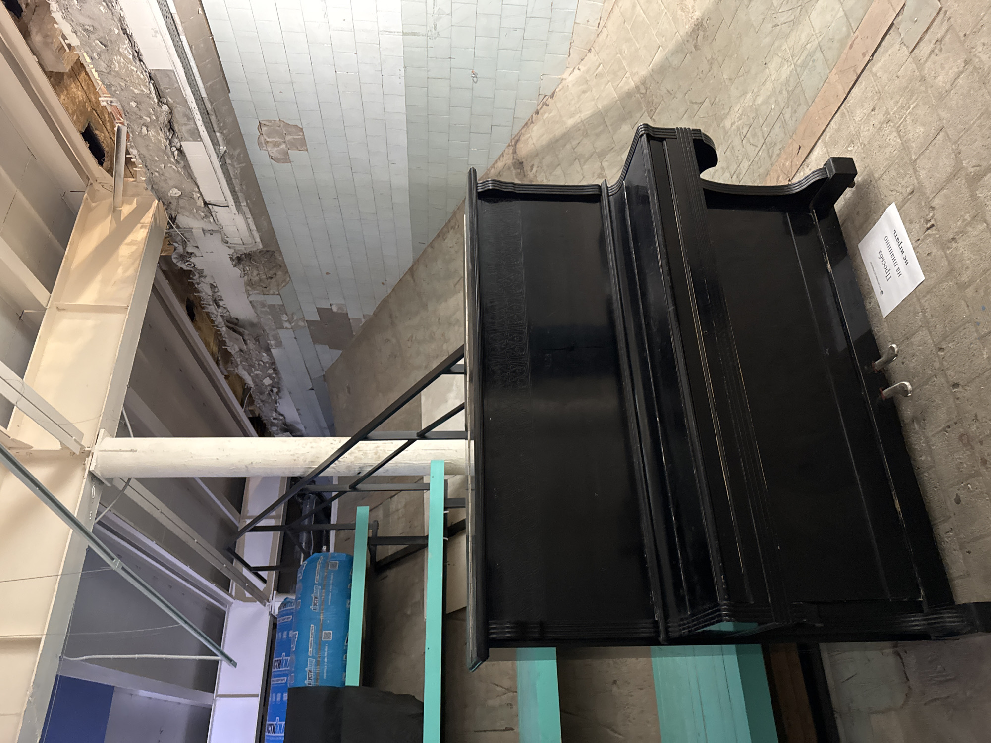 The image shows an interior under renovation. The perspective is unconventional, possibly taken from a low angle or with a distorted lens.
On the right side, a black, possibly antique, dresser or cabinet stands against a roughly finished concrete wall. A piece of paper with text in Russian ("Просьба на машшно не шпирять," likely a request to not lean on the cabinet) is attached to the wall next to it.
Above, the ceiling has a section of pale blue-tiled area transitioning into a rougher, unfinished section with exposed brick and remnants of a former ceiling.
To the left, there's construction framing made of white painted steel beams, indicating ongoing work. Blue-wrapped bundles and turquoise painted wooden pieces are scattered nearby.
Based on the style of the building and the signage in Russian, the location is likely in Saint Petersburg, Russia. The building appears old and is in the process of being renovated or restored. No people are visible in the image.