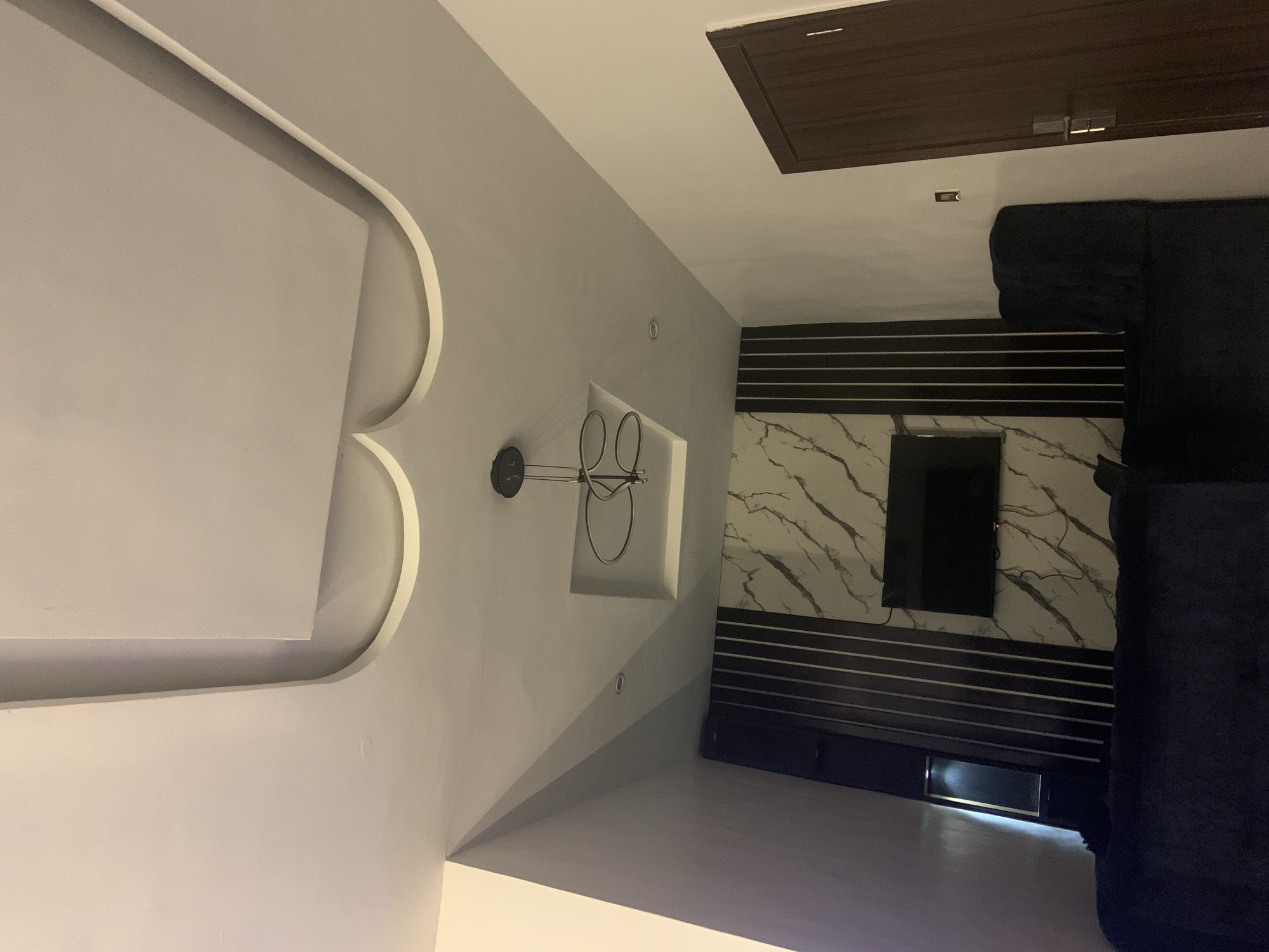 The image depicts an interior space. The ceiling features a design with curved recessed elements, and a brown access panel. A circular light fixture hangs on the wall. The wall behind a television is decorated with horizontal black stripes alternating with a marble-like pattern. A dark blue sofa partially occupies the right side of the frame. Based on the user's prompt about Ota, Nigeria, it is inferred that this is the location, although no specific identifying features are visible in the image. There are no people visible.