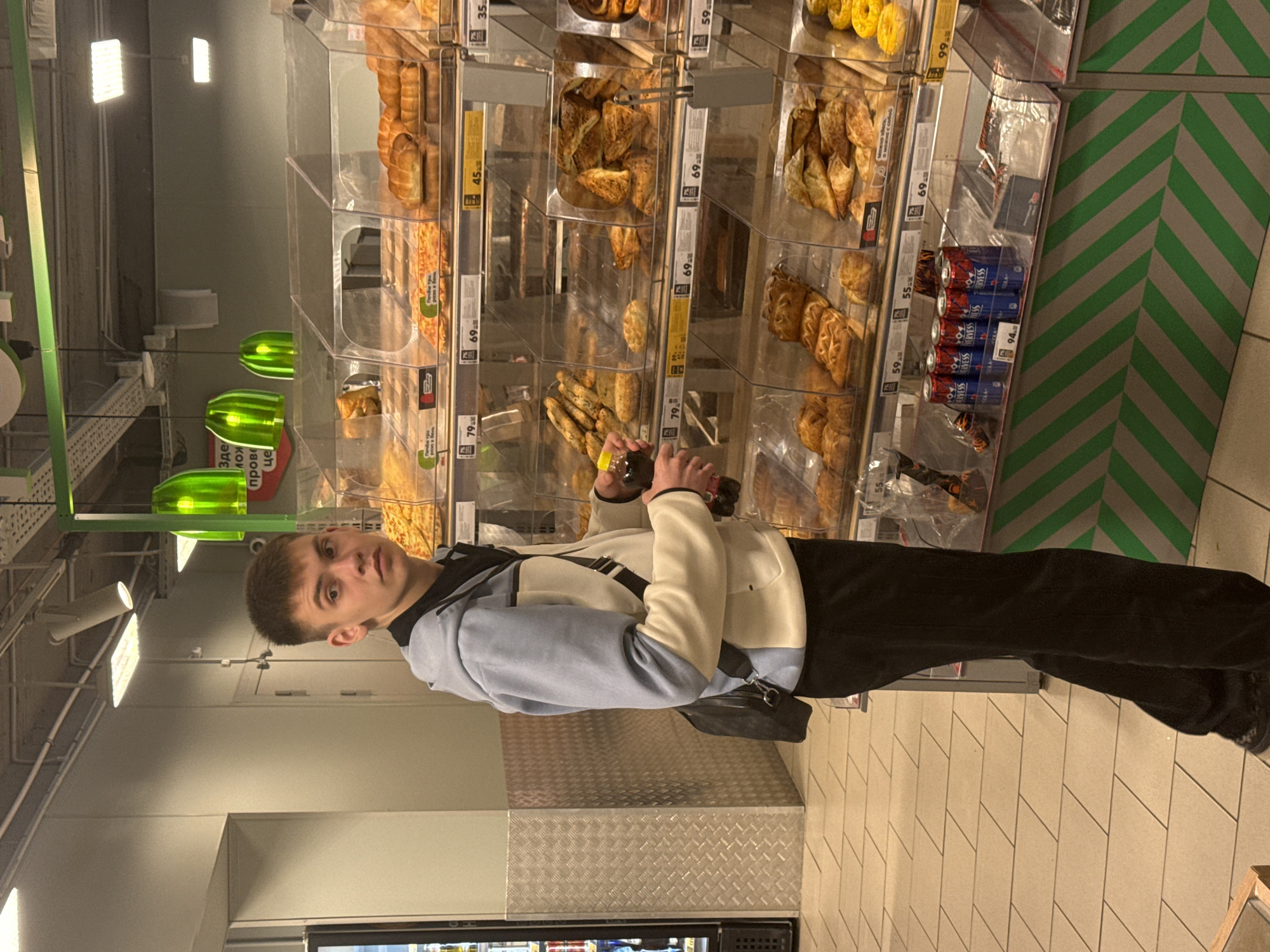 The image shows a young man standing in a grocery store. He has short, dark hair and is wearing a light blue and white jacket with black pants. He is holding a bottle in his hands. Behind him is a display case filled with pastries and bread. Price tags are visible on the shelves. The environment is a typical grocery store aisle with fluorescent lighting and tiled floors. The text "Здесь пров... це" (Here pro...) indicates the location is in a Russian-speaking area, possibly Birobidzhan, Russia.
