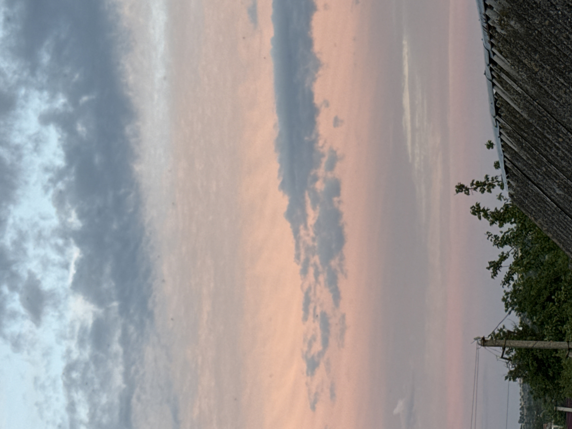 The image depicts a sky with scattered clouds in hues of blue, gray, and pink, indicative of either sunrise or sunset. Below the sky, the upper portion of a dark, corrugated roof is visible, angled from the bottom right corner upwards. To the left of the roof, several green trees and shrubs are present. A concrete utility pole with wires is also visible amongst the foliage. The background shows a distant, undulating landscape with more trees and what appear to be structures, suggesting a rural or suburban setting outside of a major city. The location is specified as Unknown, Unknown.