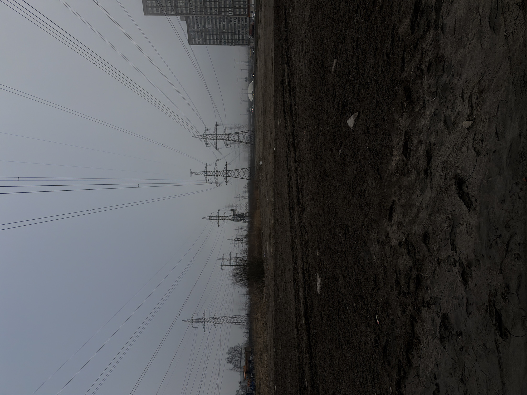The image presents a landscape in Murino, Russia under a cloudy, overcast sky. The ground is dark, possibly muddy, with visible footprints and scattered debris. A line of electrical transmission towers stretches into the distance, their wires intersecting the sky. In the background, a tall, modern building is visible, suggesting an urban or suburban area. No people are visible. The overall impression is one of an industrial or undeveloped area on the outskirts of a city.