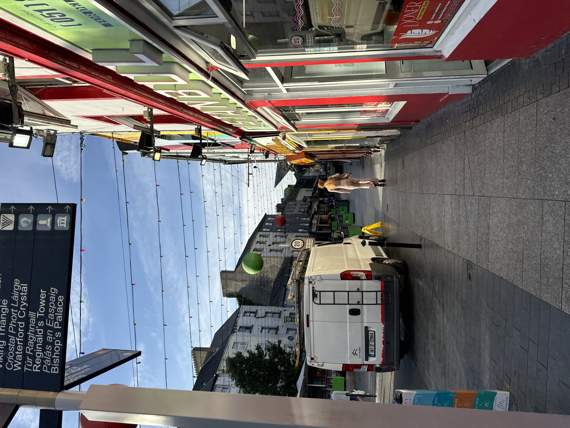 A street scene in Waterford, Ireland is captured in this full-frame, eye-level, medium-shot photograph. The image focuses on a pedestrian walkway lined with colorful buildings, suggestive of a commercial district. A white van is parked on the street on the left side of the image. String lights are suspended across the street, illuminating the scene. A person in a light-colored outfit is walking away from the camera on the right side of the street. The background shows traditional Irish architecture, with buildings of varying heights and styles. The sky is a clear blue with a few wisps of clouds.