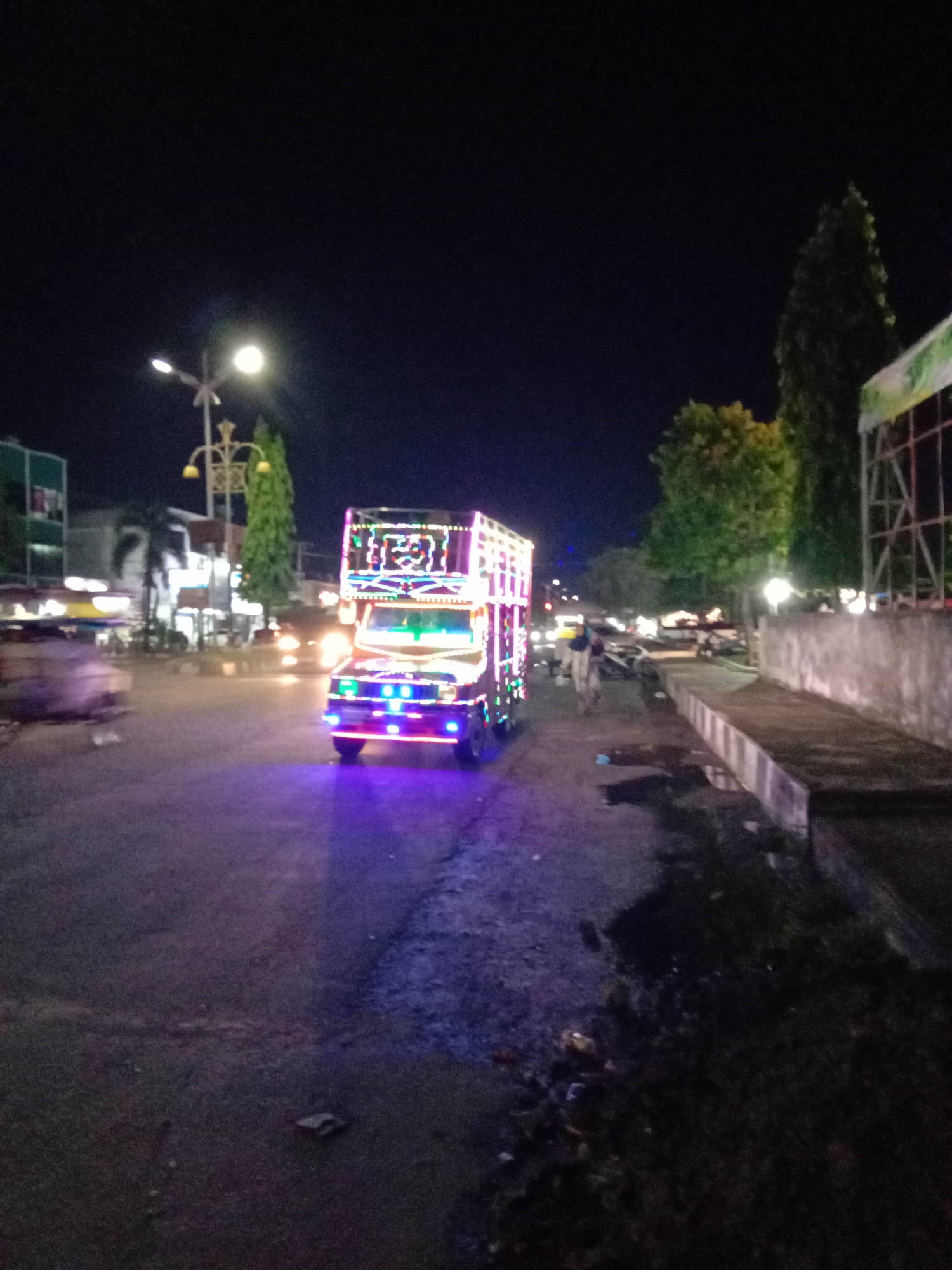 The image shows a truck brightly decorated with lights driving on a dark street at night in Lhokseumawe, Indonesia. The truck is moving towards the camera. A street lamp illuminates the area, with buildings and trees visible in the background. A person is walking along the sidewalk to the right of the truck. A concrete structure runs along the side of the sidewalk. The road surface is uneven.