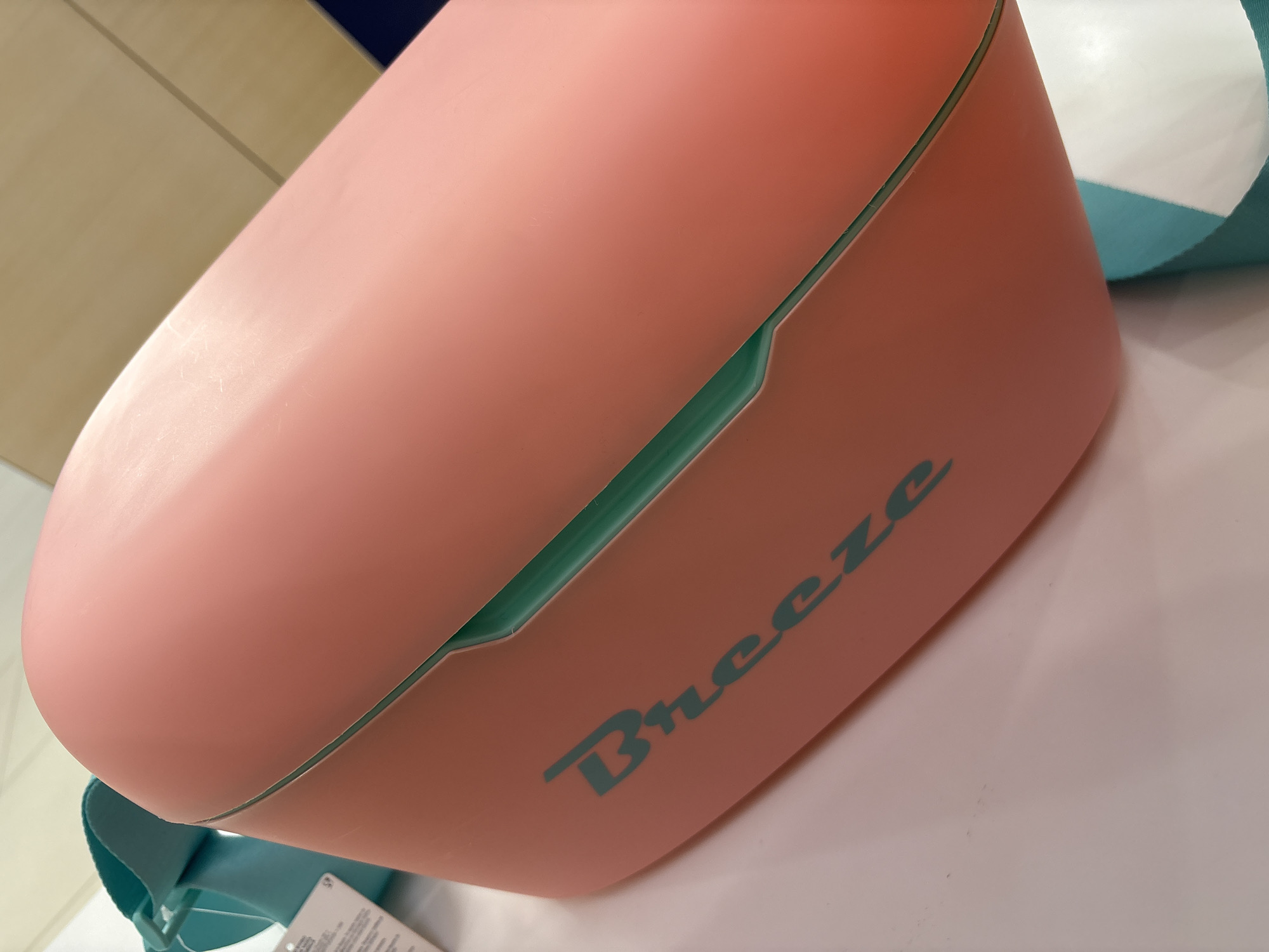 The image shows a pink container with the word "Breeze" printed on its side in turquoise. The container appears to be a rectangular prism with a curved top. The lid has a recessed edge, revealing the turquoise color of the lower section. The container sits on a white surface. The background is blurred, but a cabinet and a cushion with white and turquoise stripes are visible. The location is not discernible from the image.