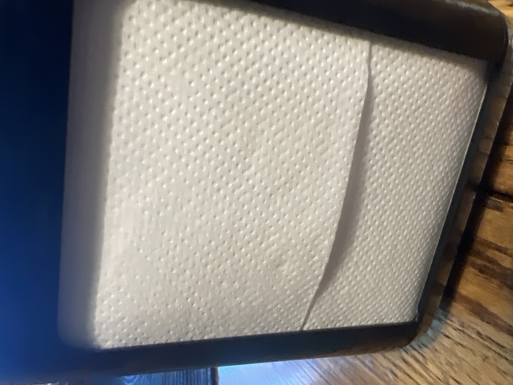 The image shows a close-up view of a napkin holder filled with white paper napkins. The napkin holder appears to be made of dark material, possibly wood or plastic, and is placed on a wooden table with a visible grain. The napkins have a textured surface with small embossed dots. A slight shadow is cast on the napkins from the top of the holder. The background is dark and out of focus, with a hint of blue. The scene is indoors, on a table likely in a restaurant or dining setting. The image does not contain any people or discernible actions. There is no information within the image to suggest a specific location in Berehove, Ukraine.