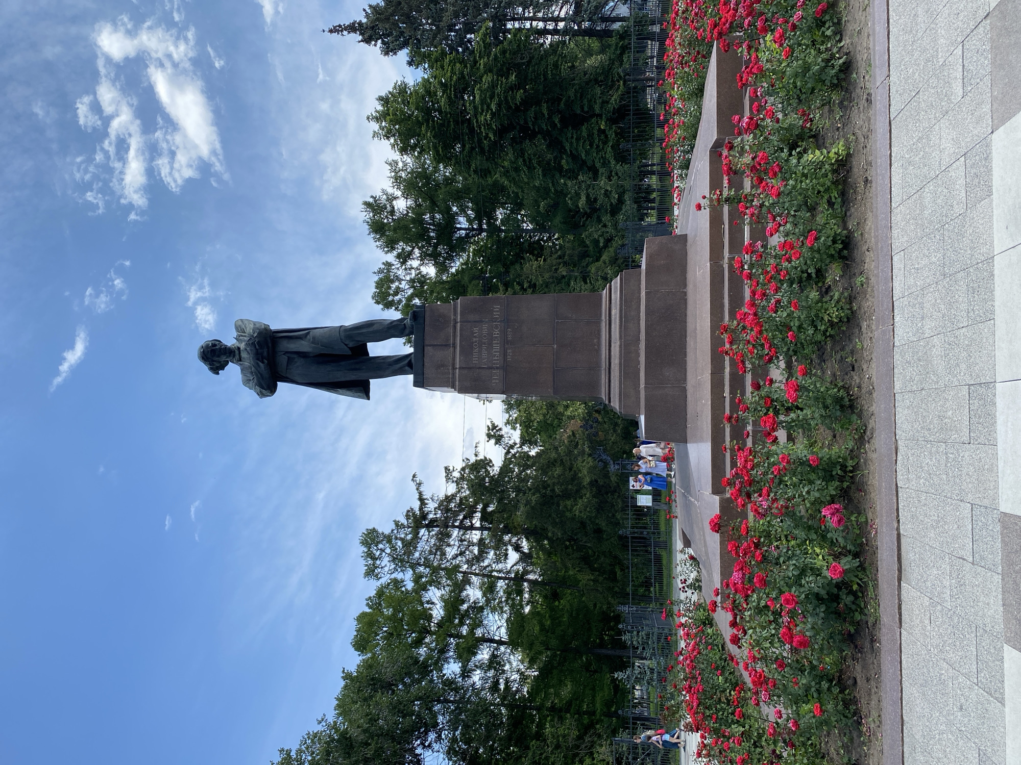 In an outdoor plaza, a bronze statue of a man in a suit and overcoat stands on a granite pedestal. The pedestal has inscriptions in Russian. A dense bed of red roses surrounds the base of the pedestal. In the background, tall green trees form a backdrop, and a black metal fence is visible. A few people are walking near the fence and in front of the trees. The sky above is blue with scattered white clouds. The foreground features a paved walkway with a grey, speckled pattern.