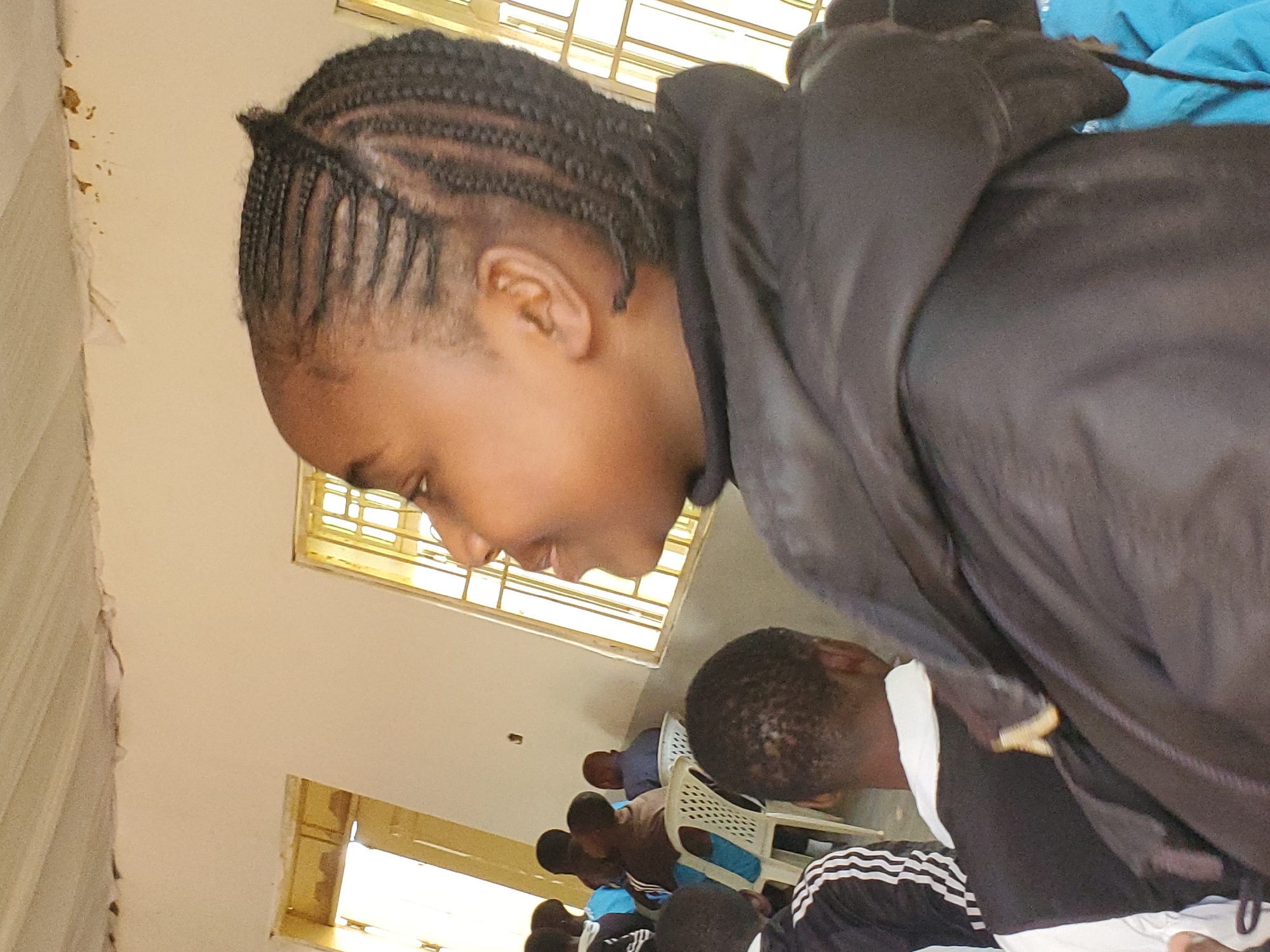 In a room with beige walls and large barred windows, several young people are seated. The camera is positioned at a low angle, focusing on the face of a young person in the foreground who is looking down and to the side, with their hair in braids. This individual is wearing a dark hooded garment that obscures their body and is partially covering another person. In the background, other individuals are visible, also looking down, suggesting they are engaged in an activity that requires their attention. Some are seated on white plastic chairs. The lighting appears to be natural, coming from the windows, and suggests it could be daytime. The overall scene appears to be an indoor gathering or educational setting in Kuru Karama, Nigeria.