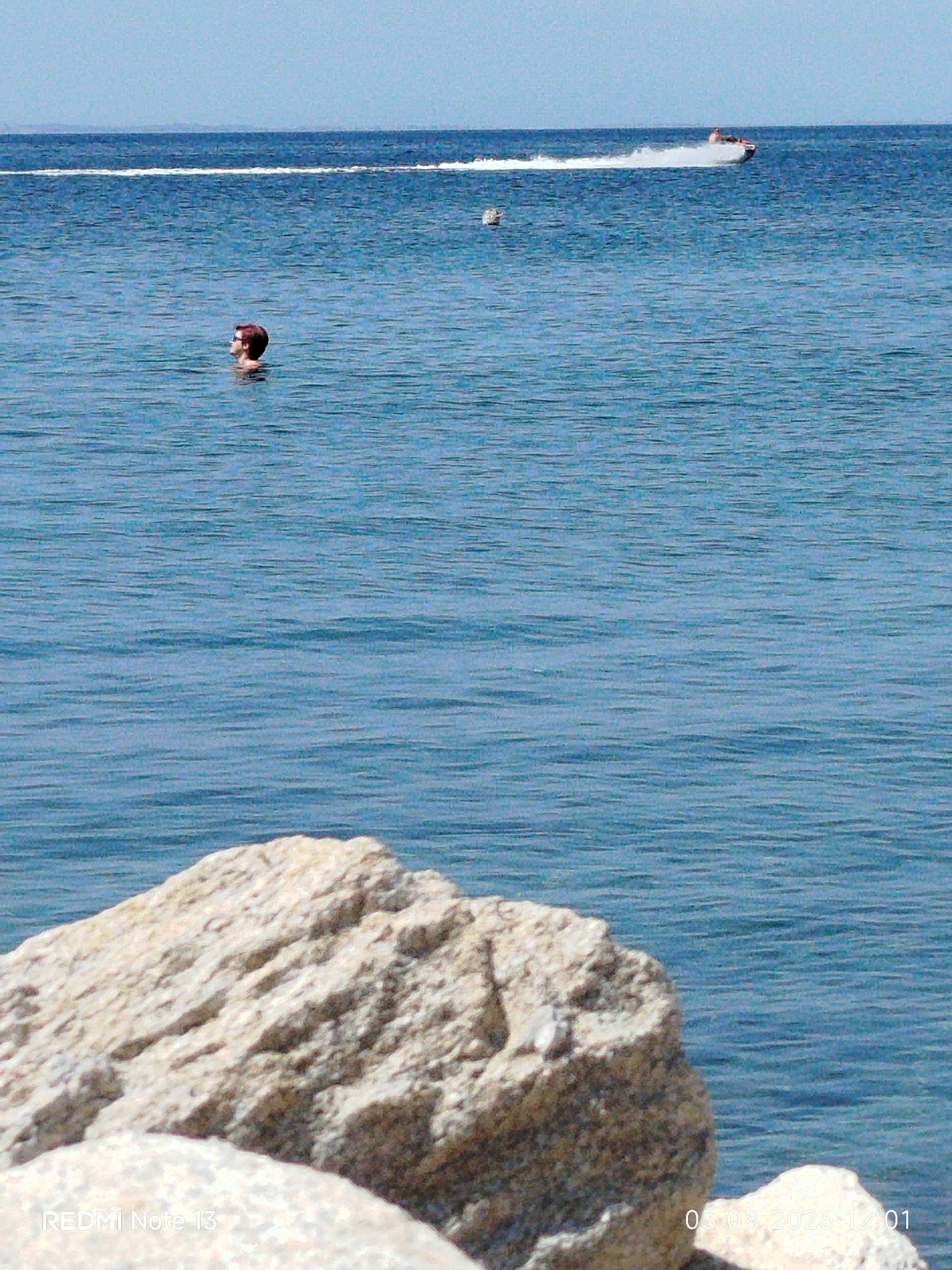 A person with short, reddish hair and glasses is swimming in the blue sea. They are looking to the left and are partially submerged. In the background, a jet ski is moving from left to right, creating a white wake. There is a white buoy or marker in the water to the left of the jet ski. In the foreground, a large, light-colored rock is visible, partially obscuring the view of the water. The sky is a clear, light blue. The timestamp "03.08.2026 12:01" and "REDMI Note 13" are visible in the bottom right and left corners respectively, indicating the image was taken on August 3rd, 2026, at 12:01 PM, with a Redmi Note 13 phone. This scene is consistent with a coastal area in Sithonia Municipal Unit, Greece.