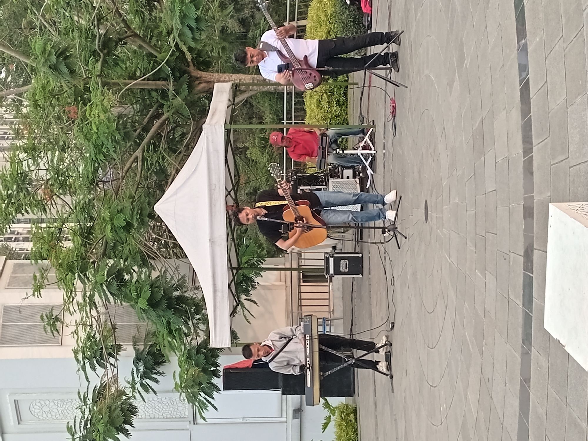 A band is performing outdoors under a white canopy tent in what appears to be a paved public space, possibly in Gunung Putri, Indonesia. The band consists of four members. On the far left, a person in a light gray hoodie and black pants plays a keyboard on a stand. To the right of the keyboardist, a singer in a black t-shirt and blue jeans plays an acoustic guitar and sings into a microphone. In the center, a person wearing a red shirt and blue jeans plays a keyboard while seated. On the far right, a person in a white t-shirt and black pants plays a bass guitar. In the background, there are trees, a building with windows, and a metal railing. The ground is paved with circular patterns and square tiles. There are speakers, cables, and a drum set visible on stage.