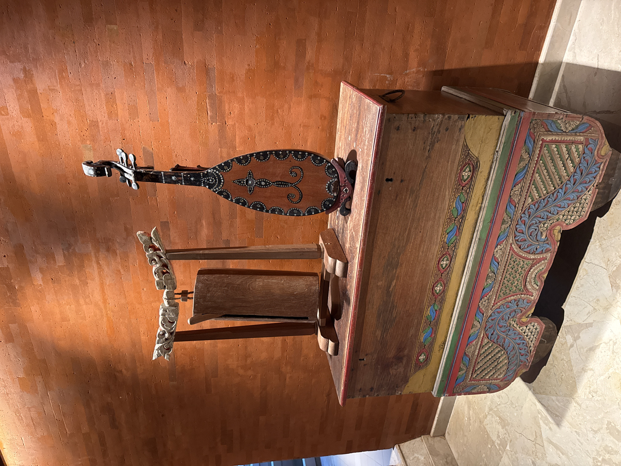 An image of a decorative wooden musical instrument with an oval body, a wooden stand, and an ornate wooden chest, photographed indoors against a brick wall. The location may be Kuta Selatan, Indonesia. The musical instrument is embellished with pearl-like decorations and rests on a wooden stand. The wooden chest is painted with colorful patterns. The room is brightly lit, highlighting the textures and patterns of the objects.