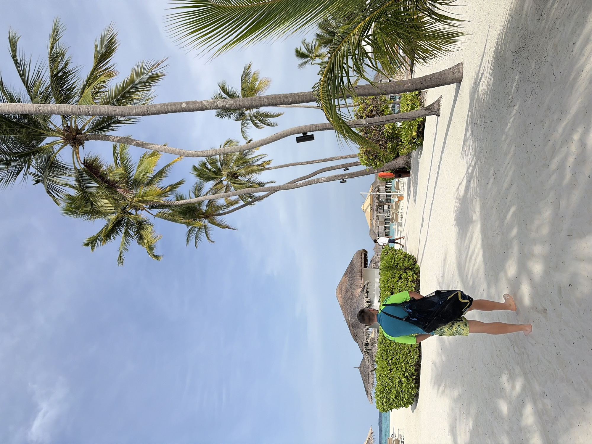 In Halaveli, Maldives, a man walks barefoot on a sandy beach carrying a black duffel bag over his shoulder. He is wearing a teal and lime green t-shirt and patterned yellow and green shorts. Palm trees with lush green fronds line the beach, casting shadows on the white sand. In the background, thatched-roof bungalows and lounge chairs are visible, suggesting a resort setting. The sky is clear and blue with a few wispy clouds. The scene depicts a serene tropical vacation atmosphere.