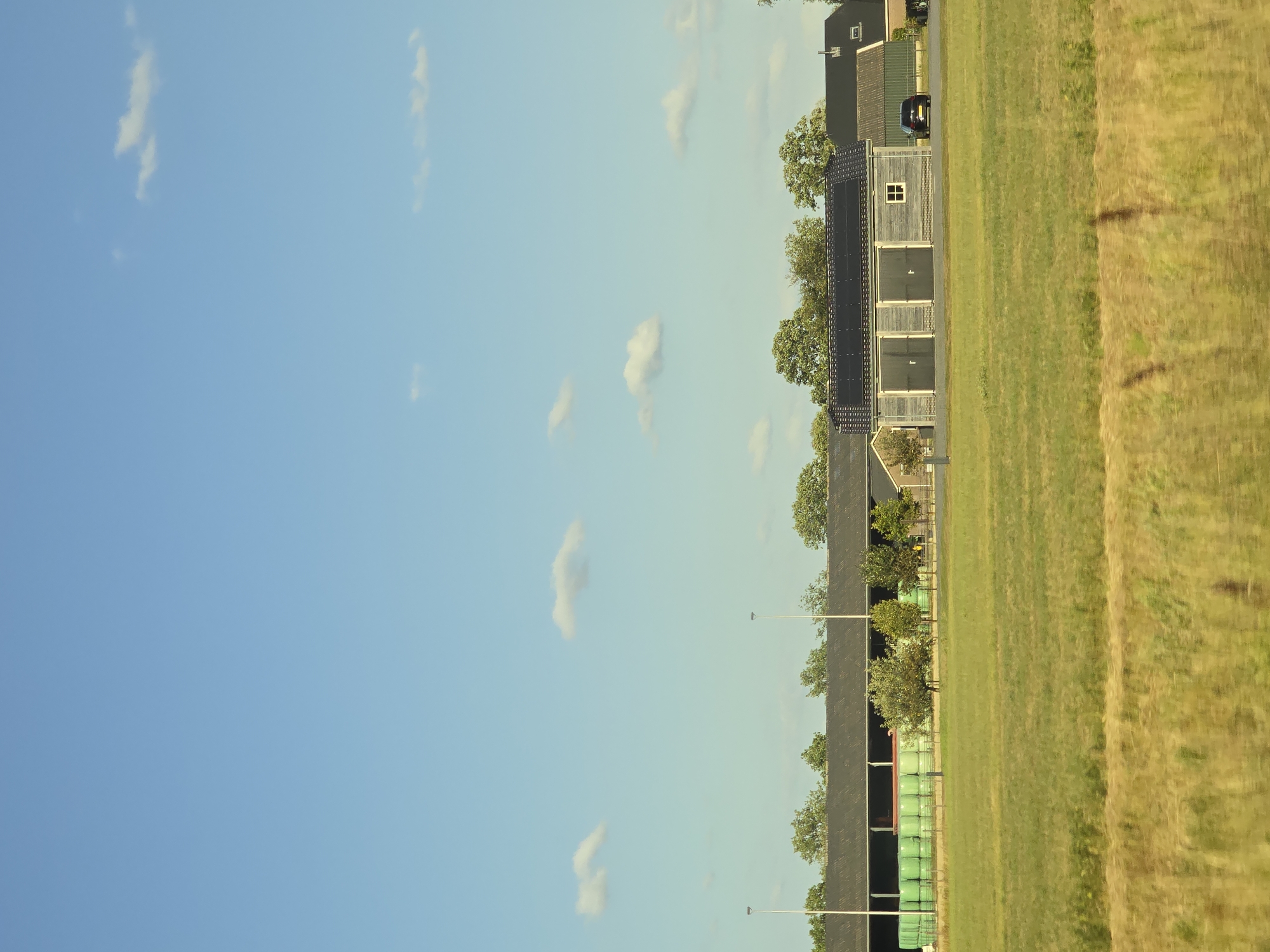 The image depicts a rural landscape under a clear blue sky dotted with a few scattered white clouds. In the mid-ground, a long, dark-roofed building with a row of evenly spaced lights extends across the frame. To the left of this building, a large collection of bright green cylindrical bales is stacked. Several deciduous trees are visible behind the building, and a smaller, wooden structure with two large doors and a small window is situated to the right, with a dark car parked in front of it. The foreground is dominated by a field of green grass, with a strip of dry, golden-brown grass in the immediate foreground, suggesting a transition in vegetation or a path. The overall scene conveys a sense of open space and agricultural activity. The location is Klarenbeek, Netherlands.