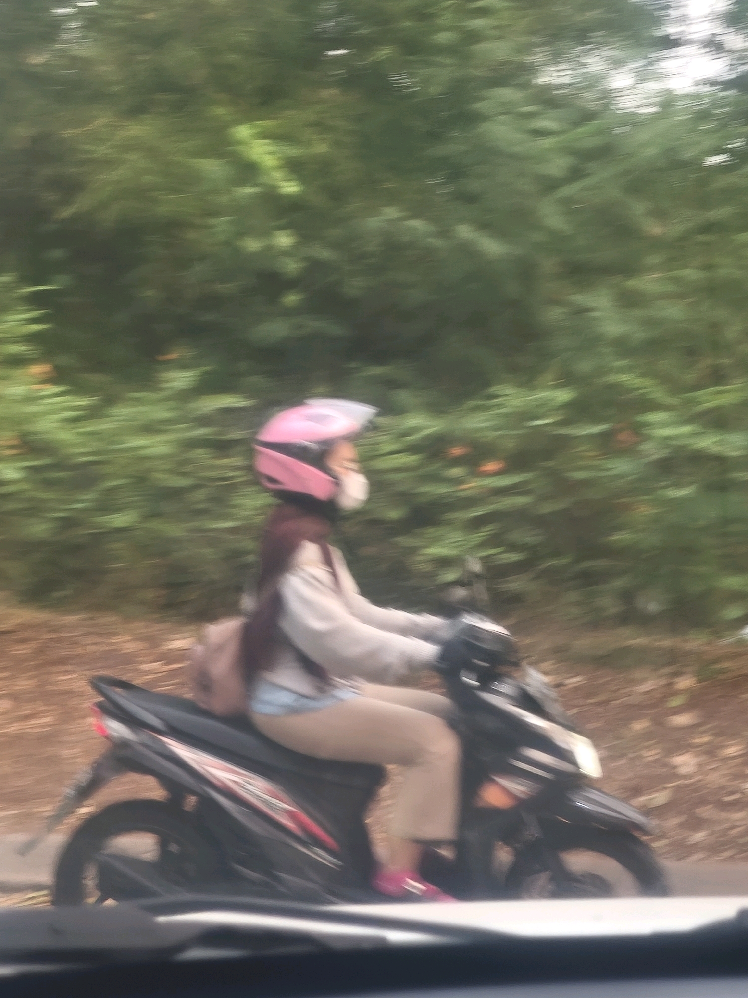 A person is riding a motorcycle on a paved road with trees on the right side of the frame. The person is wearing a pink helmet and a light-colored jacket. The motorcycle is black and has red accents. The scene appears to be in motion, as indicated by the motion blur in the image. The location is the city of Karangsatria, Indonesia.