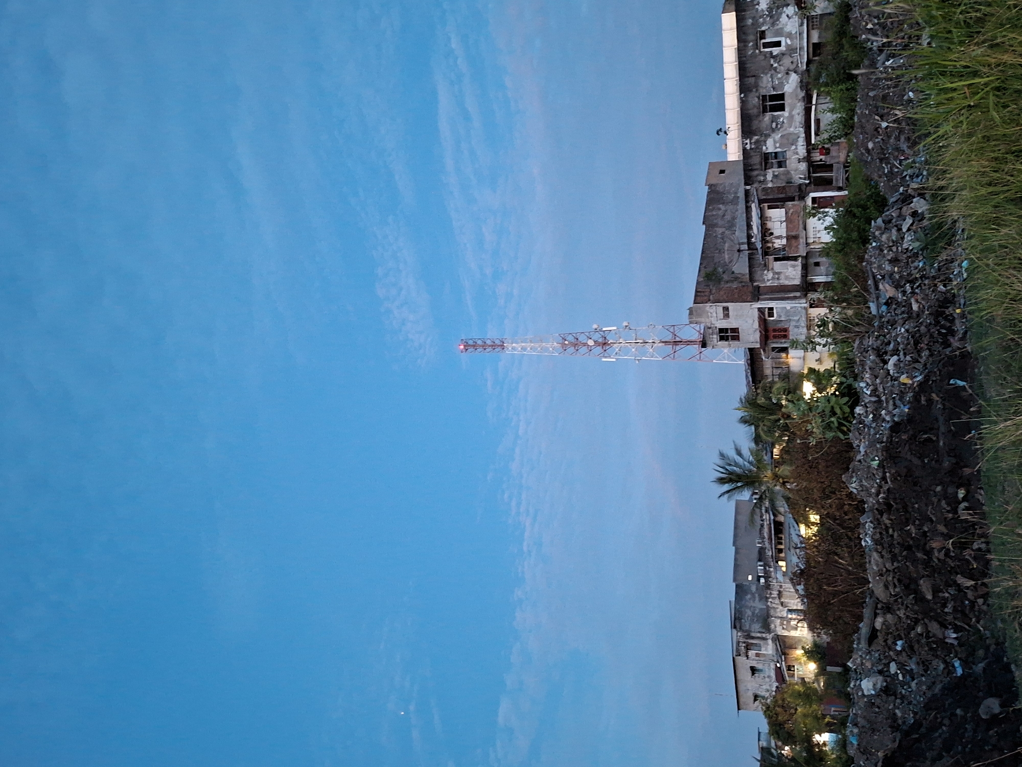 The image depicts a tower adjacent to buildings and trash. The sky is blue and somewhat cloudy. The buildings are run down. The location is the city of Lhokseumawe, Indonesia. The image is rotated by ninety degrees clockwise.