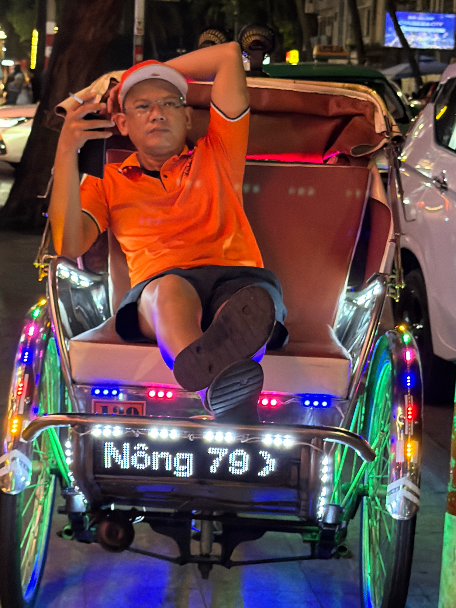 A man in an orange shirt and a red and white Santa hat sits in a brightly lit pedicab in Nha Trang, Vietnam. The pedicab is decorated with numerous colorful LED lights on its wheels, frame, and rear display. The man is resting his head on his left arm and holding a mobile phone in his right hand. The rear of the pedicab has a digital display showing the text "Nông 79J" in white LED lights. The background shows a nighttime street scene with other vehicles, including a white car and a green taxi. The overall impression is a festive and illuminated nighttime street scene in Vietnam.