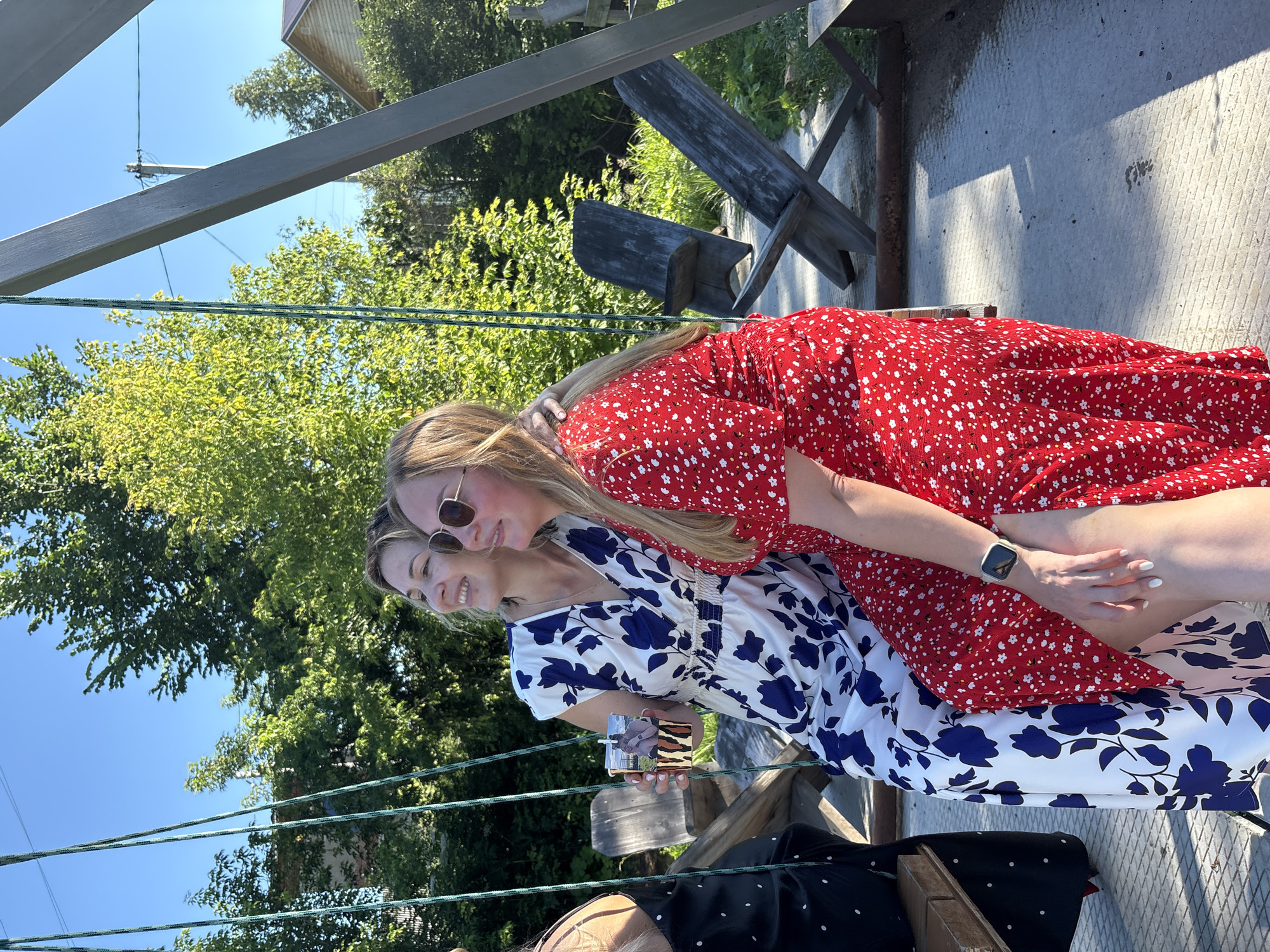 Two women are seated on a swing on a sunny day. The woman on the left wears a white dress with a blue floral pattern. She holds a cup with a photo of two people in it. The woman on the right wears a red dress with a white floral pattern and sunglasses. She has her arm around the other woman's shoulders. They are in an outdoor setting with trees and wooden chairs visible in the background. This scene is located in Zav'yalovo, Russia.