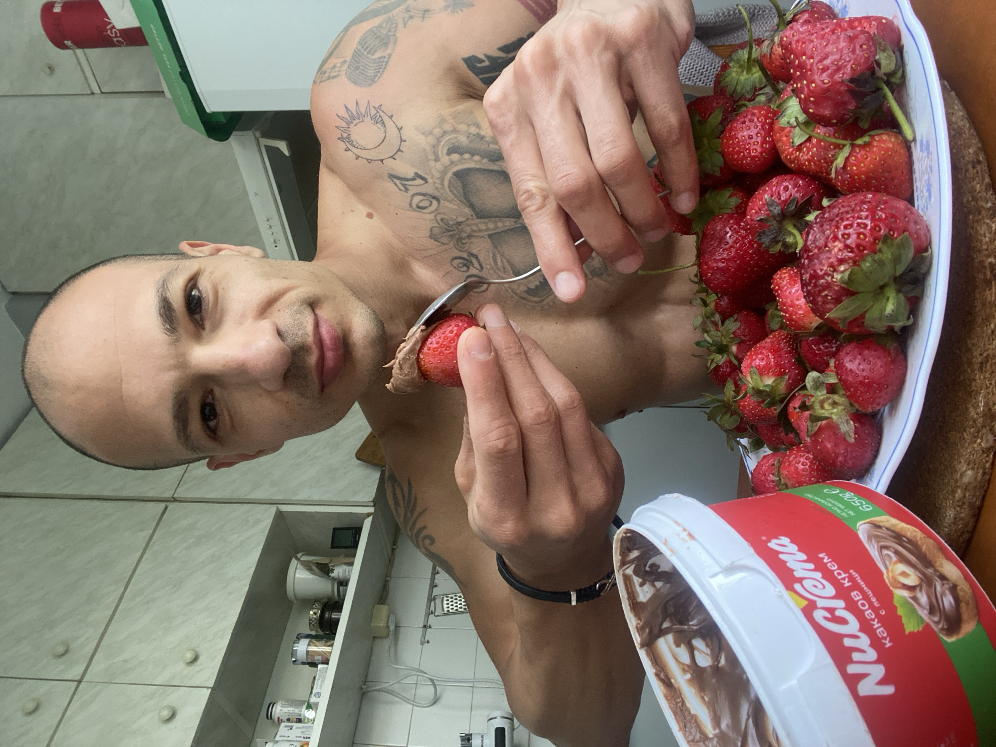 A bald, tattooed man is in a kitchen, holding a spoon with a strawberry covered in chocolate spread. A large tub of NuCrema chocolate spread is on the left, and a plate full of strawberries is on the right. The kitchen has white cabinets and shelves stocked with various items. The man is looking at the camera. The scene appears to be in Pleven, Bulgaria, as indicated by the text on the chocolate spread tub.