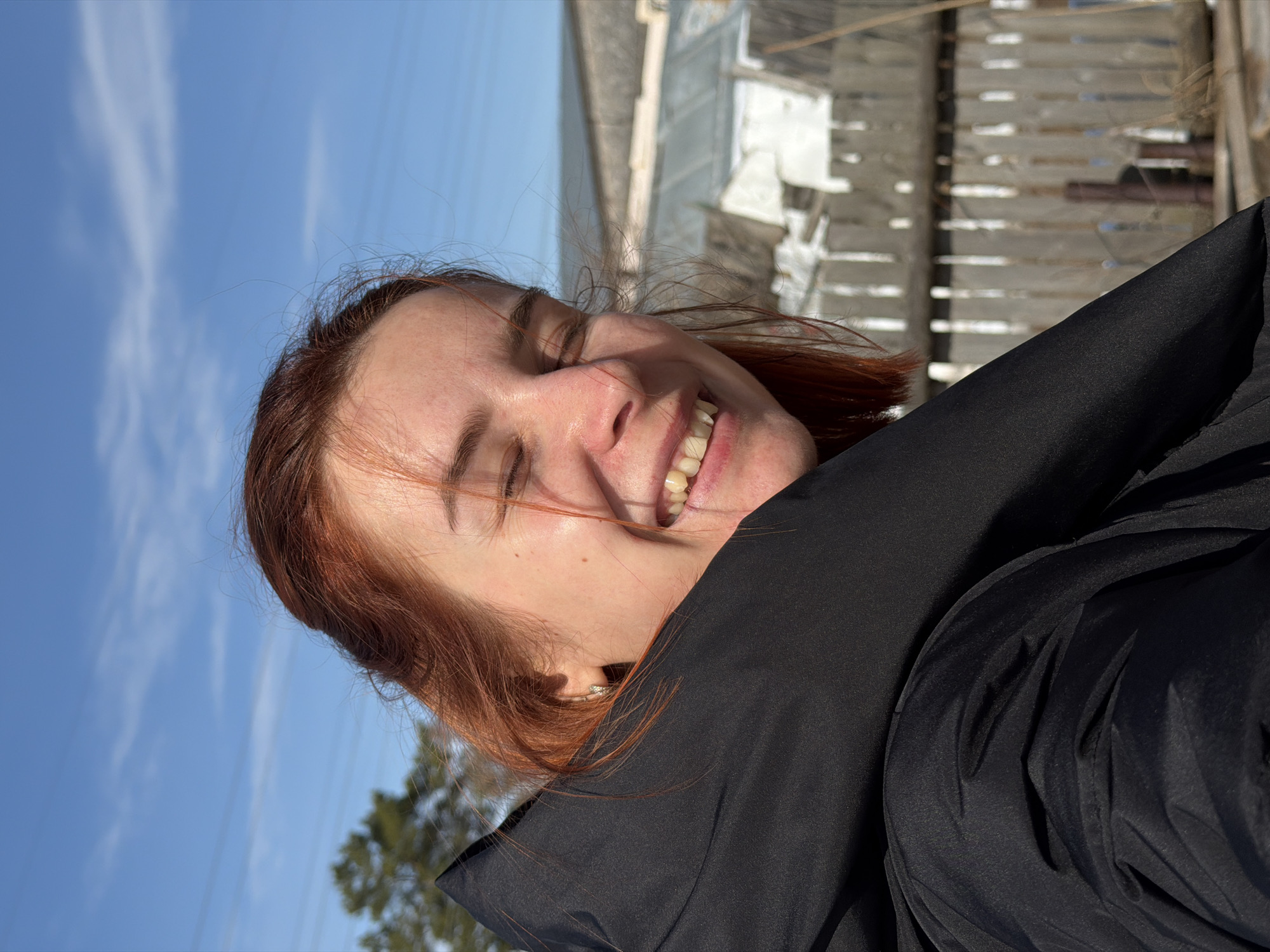 The image displays a close-up, outdoor portrait of a female subject, angled slightly to her left. Her reddish-brown hair is visible, with several strands loose around her face. Her eyes are closed, consistent with squinting in bright sunlight or a strong smile, and her mouth is open in a wide grin, revealing both upper and lower teeth. Her skin appears fair, with small moles or freckles on her right cheek. She is wearing a dark, likely black, jacket with a prominent collar that partially obscures her lower face and left shoulder. A small silver hoop earring is visible in her left earlobe. The background, largely out of focus, shows a clear blue sky with sparse, wispy clouds in the upper left. A portion of a green coniferous tree is visible on the far left. To the right, a weathered wooden picket fence stands, with snow or dry vegetation covering the ground at its base. Beyond the fence, out-of-focus structures are present, including a building with a light-colored, possibly corrugated, roof and a darker building behind it. Faint power lines are discernible against the sky in the upper right. The scene is brightly illuminated, indicating a sunny day. The location depicted is the city of Chaikovsky, Russia.