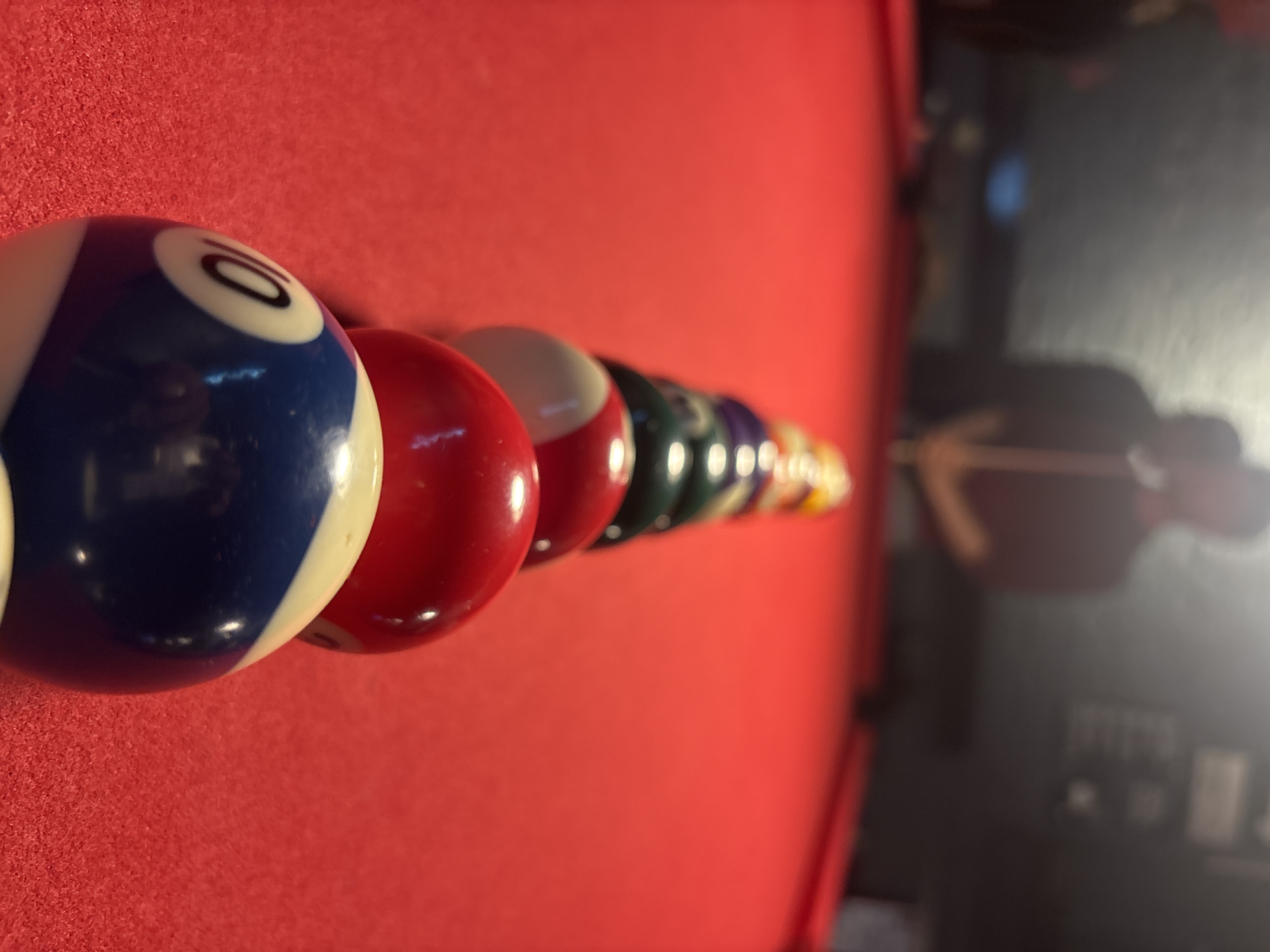 The image shows a close-up of a rack of billiard balls on a red pool table. The balls are arranged in a row, with a blue and white "10" ball in the foreground, followed by a red ball, then white and red, green, and several other balls fading into the background. The pool table is red, and the background to the right shows a blurred reflection of a person. The table's legs and possibly a bar-like area are also in the blurred background. There is no information in this image to indicate it is located in Mexico City, Mexico.