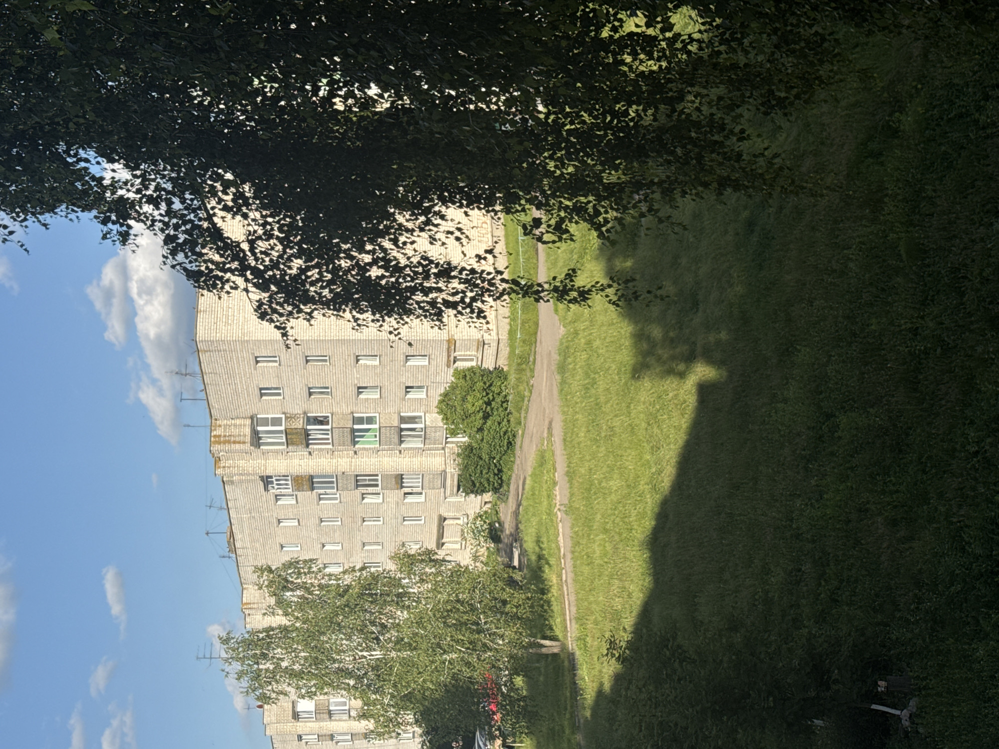 An exterior, eye-level, medium shot shows a multi-story apartment building under a cloudy blue sky. The building is made of light-colored brick and has numerous windows, some with balconies. A thin, mature tree with light bark and green leaves stands to the left of the building. In the foreground, the ground is covered in grass, with some areas in shadow. To the right, the branches of a large, leafy tree partially obscure the view of the apartment building. The setting appears to be a residential area within the Яромінскі сельскі Савет in Belarus.