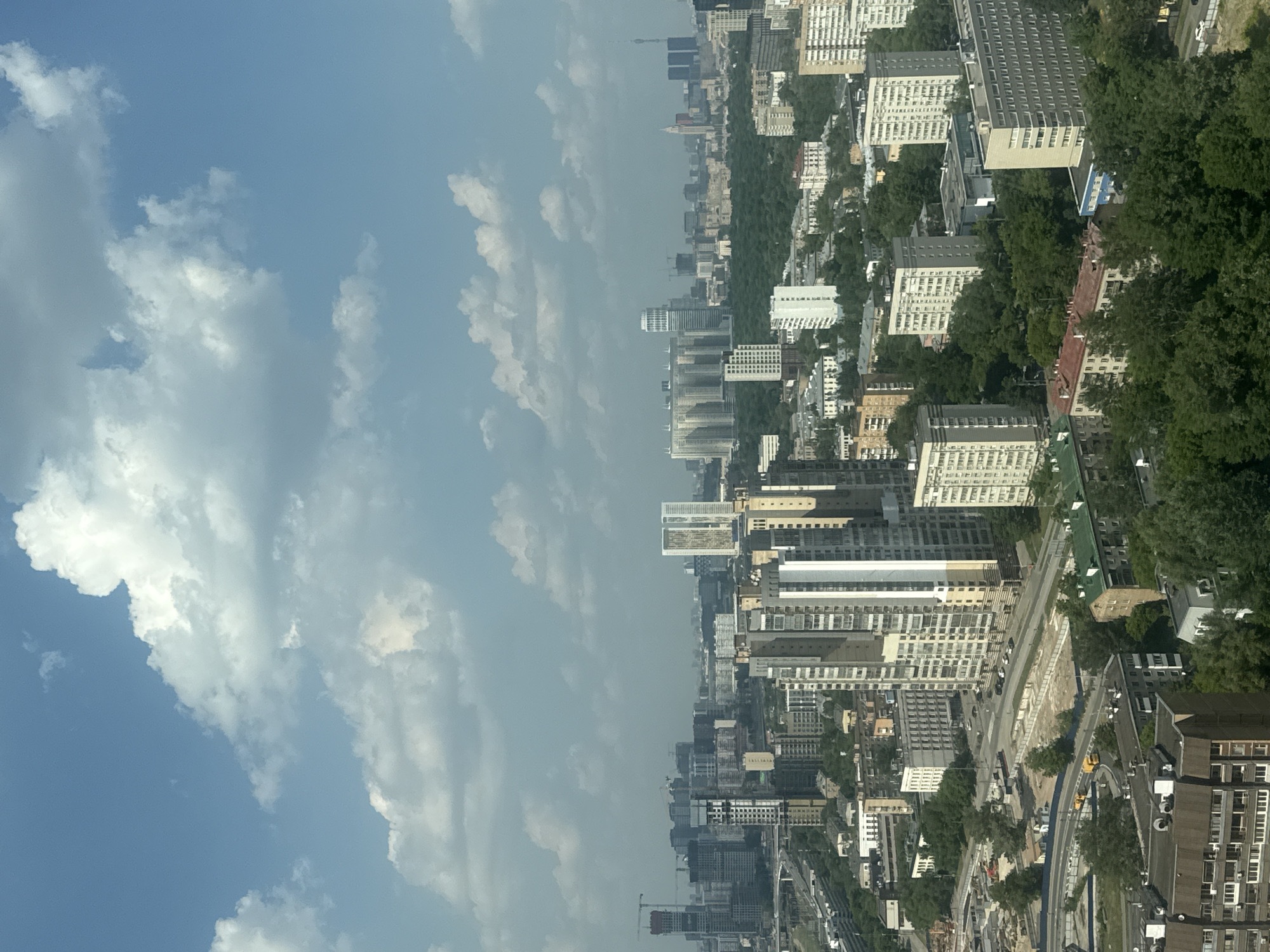 This is a high-angle, eye-level, full shot of the city of Khimki, Russia, under a bright, partly cloudy sky. The foreground is dominated by the tops of several residential buildings, some with balconies and satellite dishes. In the mid-ground, a mix of modern and older apartment buildings, office buildings, and a large park or green space are visible, interspersed with trees. Construction cranes and unfinished buildings suggest ongoing development. Roads with a few yellow buses and cars weave through the urban landscape. The background features a dense skyline of numerous high-rise buildings, including apartment complexes, office towers, and what appears to be a television tower on the far right. The overall impression is of a large, sprawling, and developing urban area.
