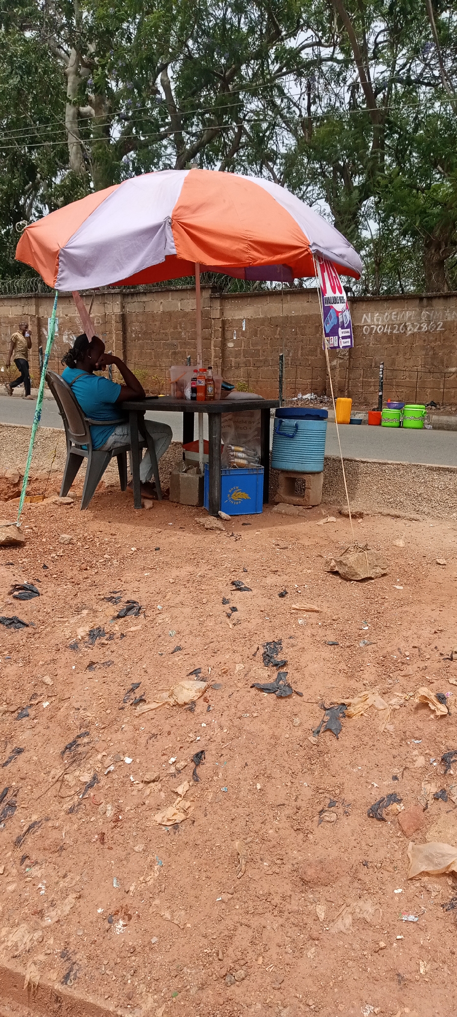 The image depicts an outdoor scene in Jos, Nigeria. A person with dark hair, wearing a blue shirt and denim shorts, is seated in a black plastic chair at a small, dark-colored table under a striped orange and white umbrella. The person has their head resting on their hand. On the table are several bottles of soda and what appears to be food. A blue plastic crate, labeled "ANDREW BREWERY" and containing bottles, is located under the table, along with other containers. A blue water cooler sits next to the table, supported by blocks.
The immediate ground area is composed of red soil littered with scattered pieces of black and clear plastic. In the background, there's a brick wall with what appears to be a small sign attached. Beyond the wall, trees and foliage are visible. There's a person walking on the sidewalk in the background. Several colorful containers are lined up against the wall. A phone number is visibly written on the wall. The scene suggests a small, informal roadside vending operation.