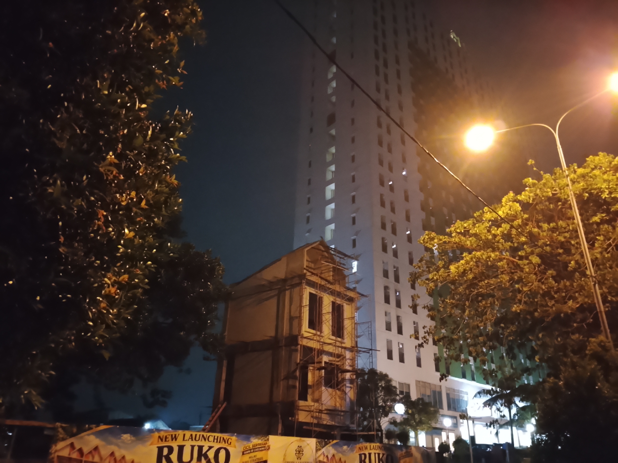 This is a nighttime, eye-level outdoor photograph taken in Depok, Indonesia, featuring a construction site in front of a modern, high-rise building. In the foreground, a banner is partially visible, displaying the word "RUKO" in large, bold lettering, indicating a commercial property development. Adjacent to this, a two-story building is under construction, surrounded by scaffolding. The building has visible window frames and a peaked roof structure. To the left and right of the frame are large trees with dense foliage, illuminated by artificial lights. The background is dominated by a tall, multi-story white building with numerous windows, suggesting it is an apartment complex or office building. Streetlights are visible, casting a warm glow and contributing to the overall nighttime ambiance. The sky is dark, indicating it is late evening or night.