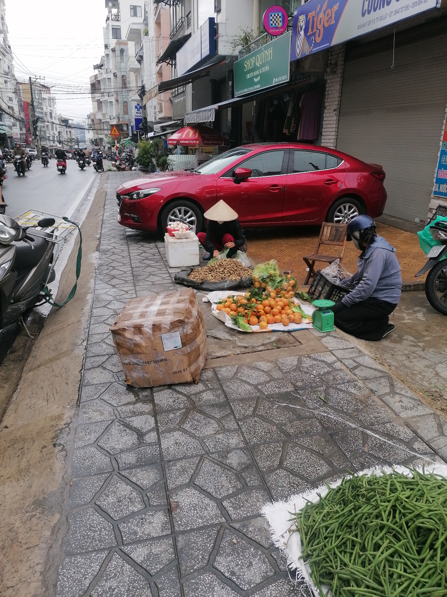 On a paved sidewalk in Da Lat, Vietnam, two vendors conduct business beside a red car parked near a street. The left vendor, wearing a conical hat, kneels next to a tray of peanuts. To the right, the second vendor, helmeted and masked, weighs produce. A bag of oranges, a scale, and crates are nearby. A large taped package sits to the left of the vendors, and a pile of green beans is in the lower right corner. Overhead, power lines crisscross above the street where motorbikes travel. Storefronts line the street, including “SHOP QUYNH” and a “Tiger” branded storefront. A street sign is visible in the distance. The OCR suggests the storefront at 06 HÀ HUY TẬP, P.3, TP. ĐÀ LẠT. The package shows 2026.01.