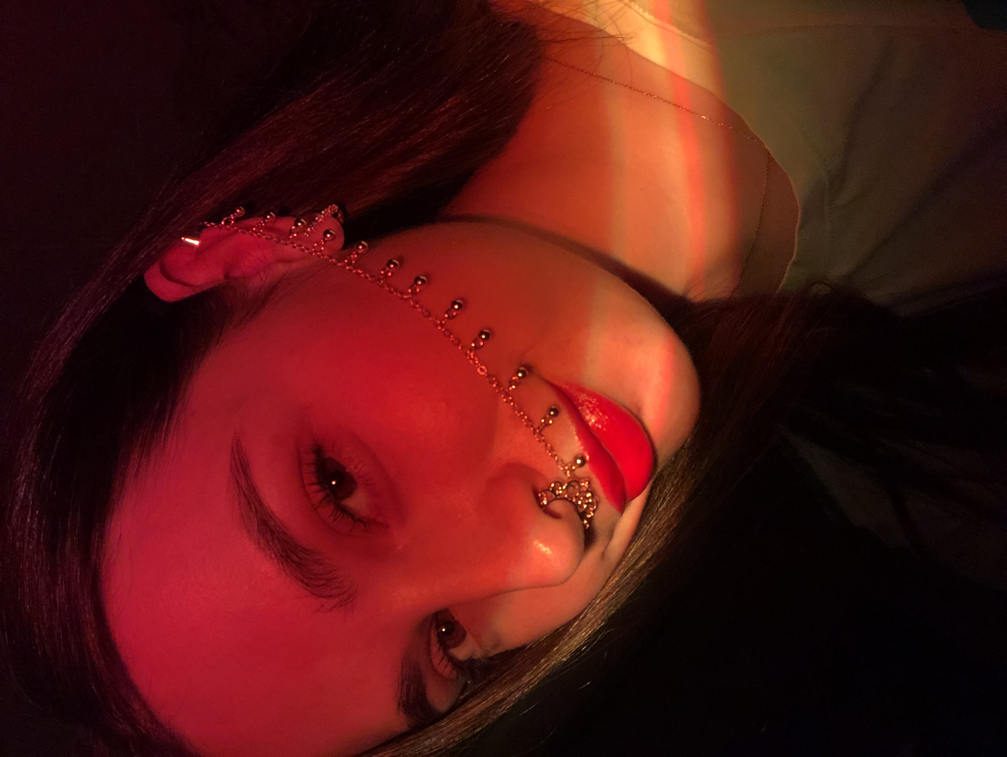 A close-up, tilted, head-and-shoulders portrait of a young woman with long, dark hair. Her face is illuminated by a predominantly red light, with additional hues of yellow and green on the upper right side of the frame. She wears a delicate gold chain headpiece that extends from her left ear, across her forehead, and down to her nose. There are multiple piercings on her nose. Her lips are painted a bright red. She also wears a very thin necklace around her neck. The background is a dark, indistinct space. The image appears to be taken indoors. No information is discernible that relates to the city of Noviy, Russia.