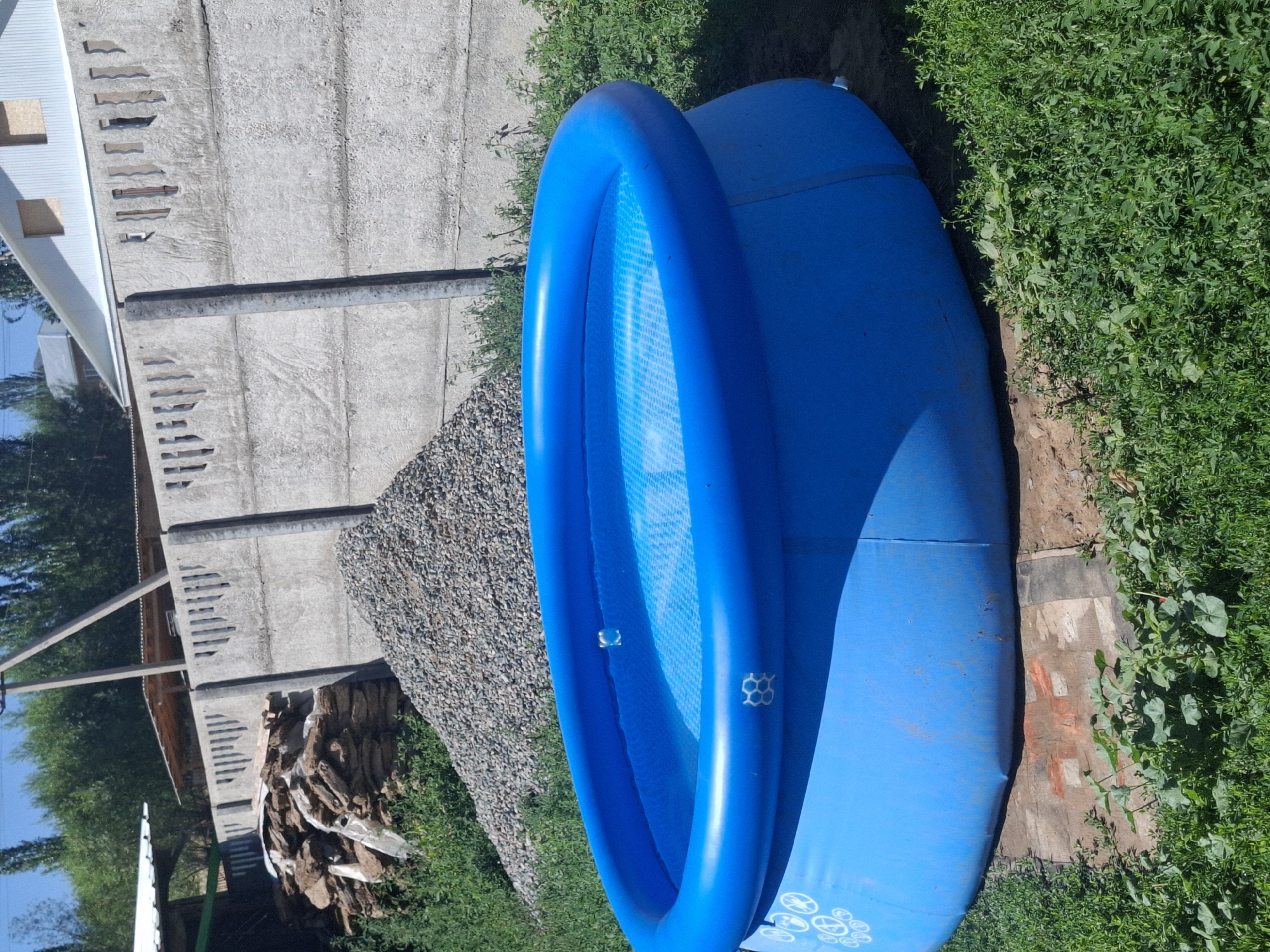 In Jeti-Ögüz, Kyrgyzstan, a bright blue, circular, inflatable pool filled with water sits on a dirt patch. The pool has white markings on its side. Behind the pool, a pile of grey gravel is visible, and further back, a low concrete fence with cutouts separates the yard from a pile of stacked stones and some wooden planks. Green grass and foliage surround the pool and the gravel pile. In the background, trees and parts of buildings, including a white-paneled house with a peaked roof, are visible under a clear sky. The scene suggests a residential area, possibly undergoing some construction or landscaping.