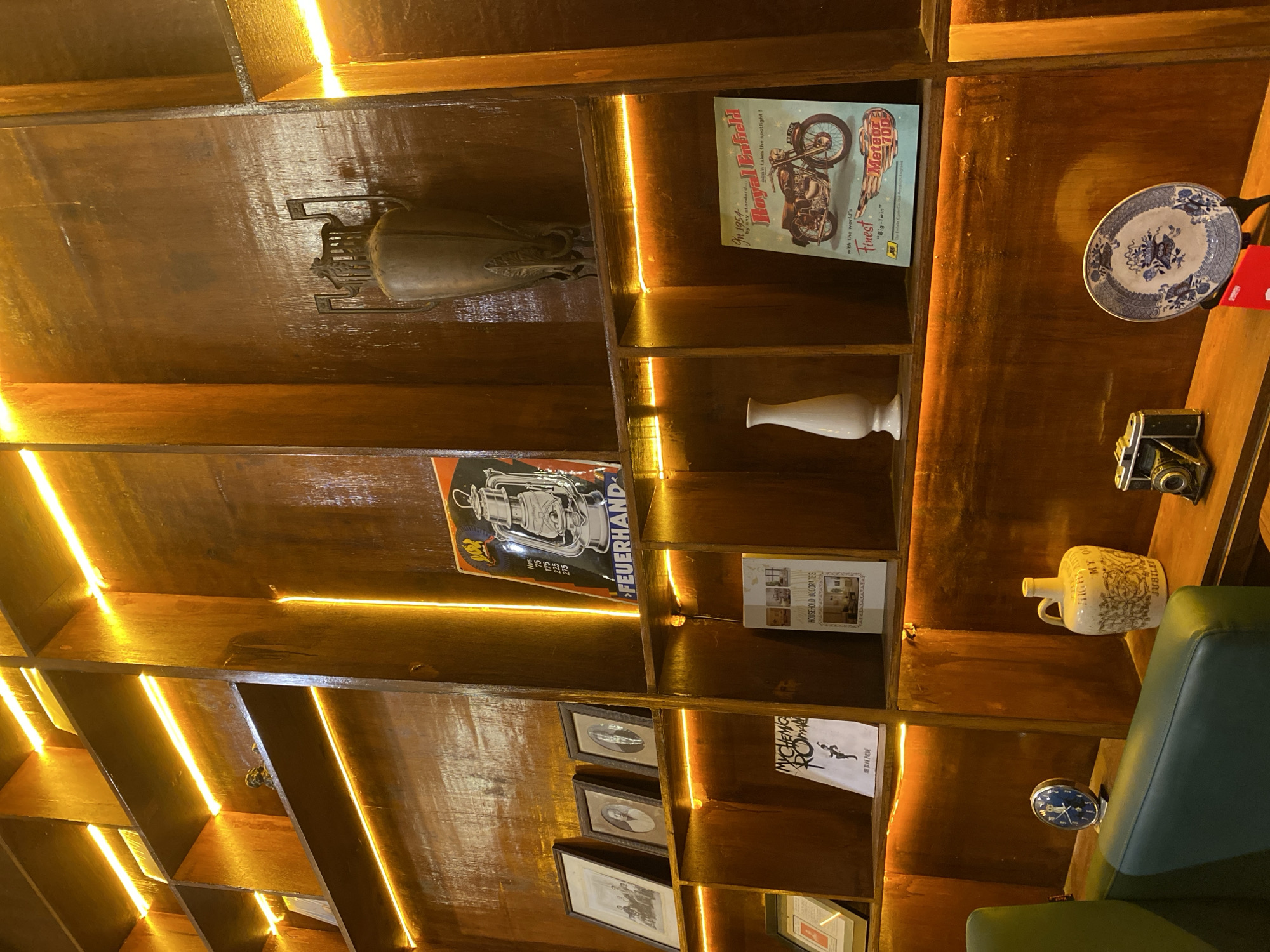 An interior shot features a tall, wooden bookshelf with multiple levels, illuminated by warm, horizontal LED strip lights. On the top shelf, a large, ornate, dark metal vase with handles stands prominently. Below this, a framed vintage advertisement for "FEUERHAND" lanterns is displayed. The next shelf down holds several framed black and white photographs. Further down, a book titled "MYCHEMIC ROMANCE" rests beside a white ceramic vase. On the right side of this shelf, a vintage poster of a Royal Enfield motorcycle, branded "Meteor 700," is visible. The bottom shelf displays a vintage alarm clock, a decorative ceramic jug with "MY QUARTZ" inscribed, a vintage camera, and a blue and white patterned plate on a stand. The overall ambiance is cozy and retro.