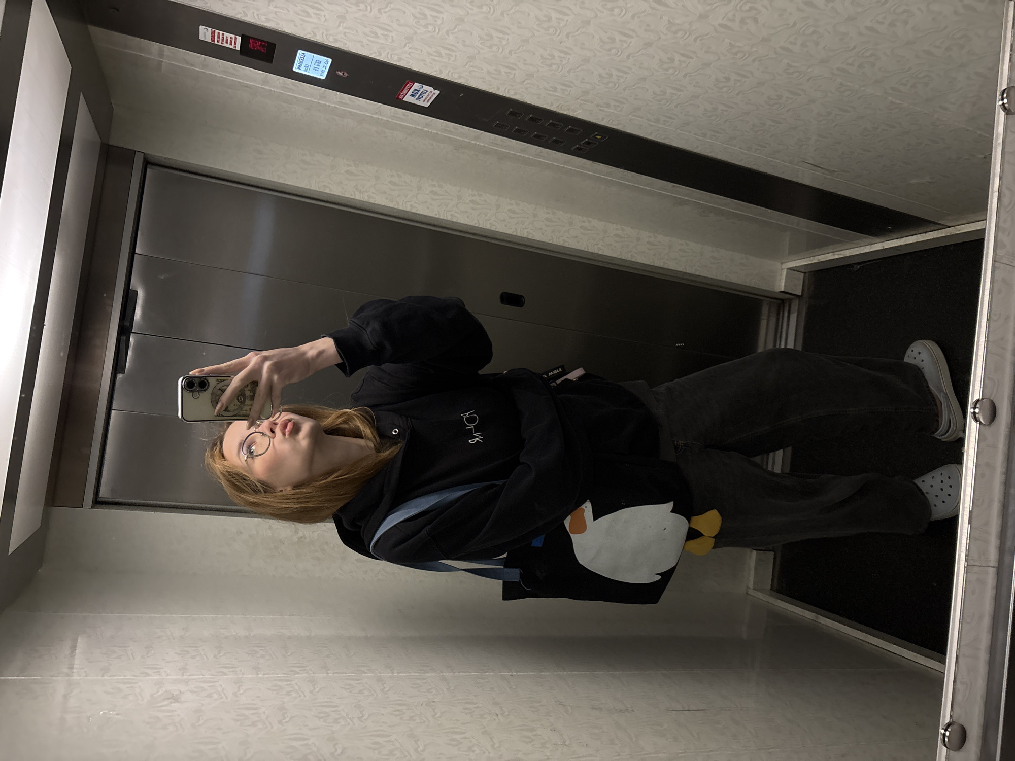 A young woman with reddish-blonde hair and glasses is taking a selfie in an elevator. She is wearing a black hoodie with a logo on the left chest, dark jeans, and light blue shoes. She is holding a smartphone in her left hand, taking the photo at roughly eye level. A backpack with a white penguin design is also visible. The elevator has a metallic door and a panel with buttons and digital floor indicators. The interior walls of the elevator are light colored. The specific location in Kragujevac, Serbia, cannot be determined from the image.