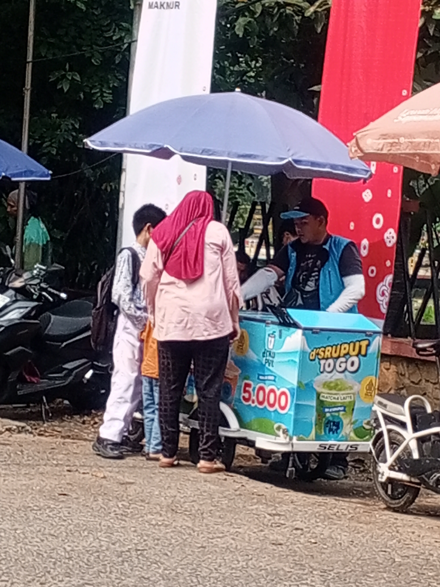 The image depicts a street scene in Depok, Indonesia, likely near a busy area. In the foreground, a person in a pink hijab and light-colored shirt stands next to a blue ice cream cart. The cart displays graphics related to "DRUPUT TO GO" and a price of "5.000." A man wearing a blue vest and a black cap is attending to the cart. To the left, two children or teenagers are visible, one wearing a backpack and the other in a patterned outfit. Several motorcycles are parked behind them, and an umbrella provides shade over part of the scene. In the background, lush green trees and vertical banners suggest an outdoor event or marketplace. The overall impression is a casual commercial interaction in a public outdoor setting within Depok.