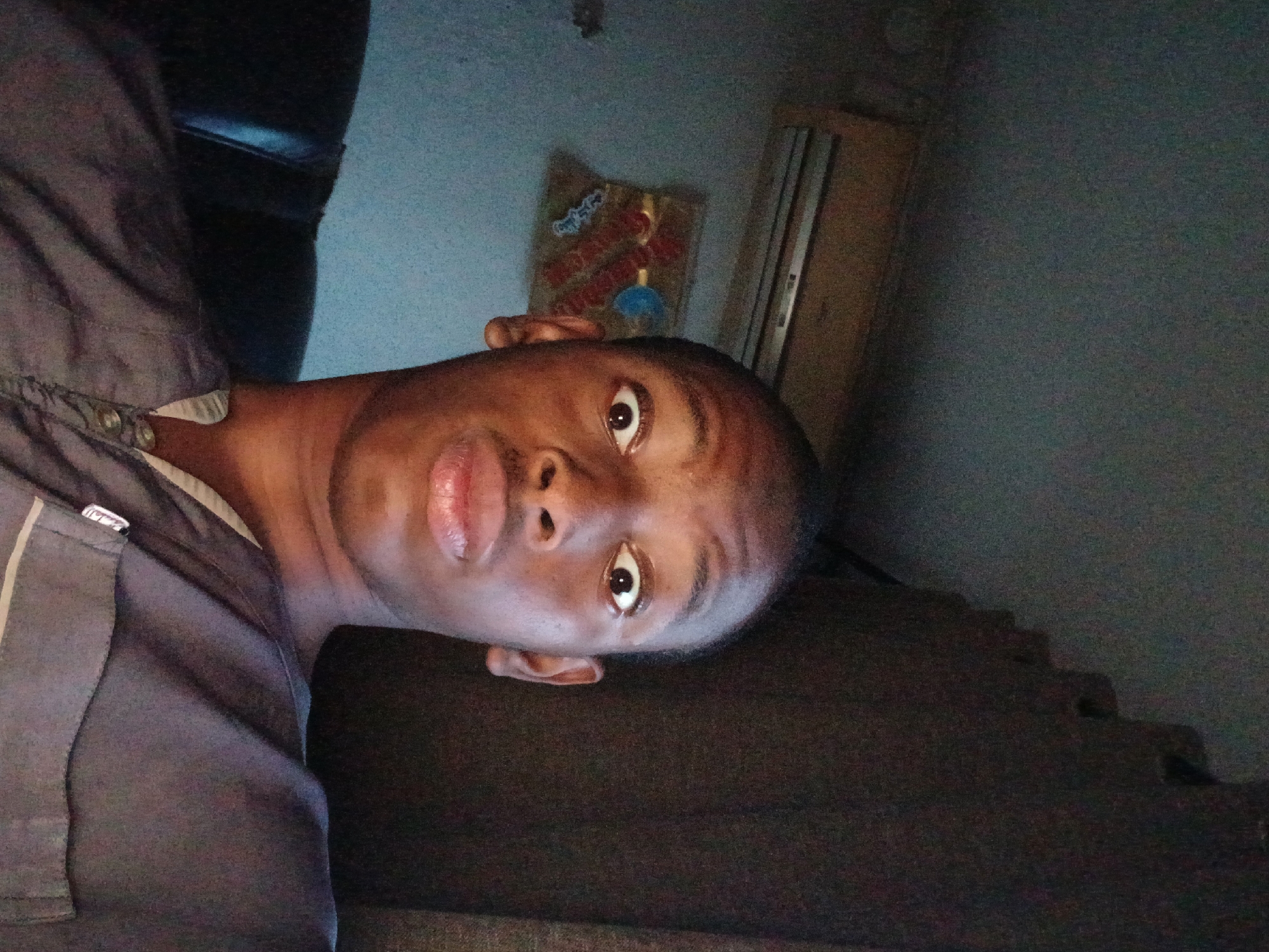 A dark-skinned person is facing the camera. The individual is wearing a gray shirt. Behind the person is a light-colored wall, a package of snacks, and a light-colored object. A dark object with steps can also be seen. The location is unknown.