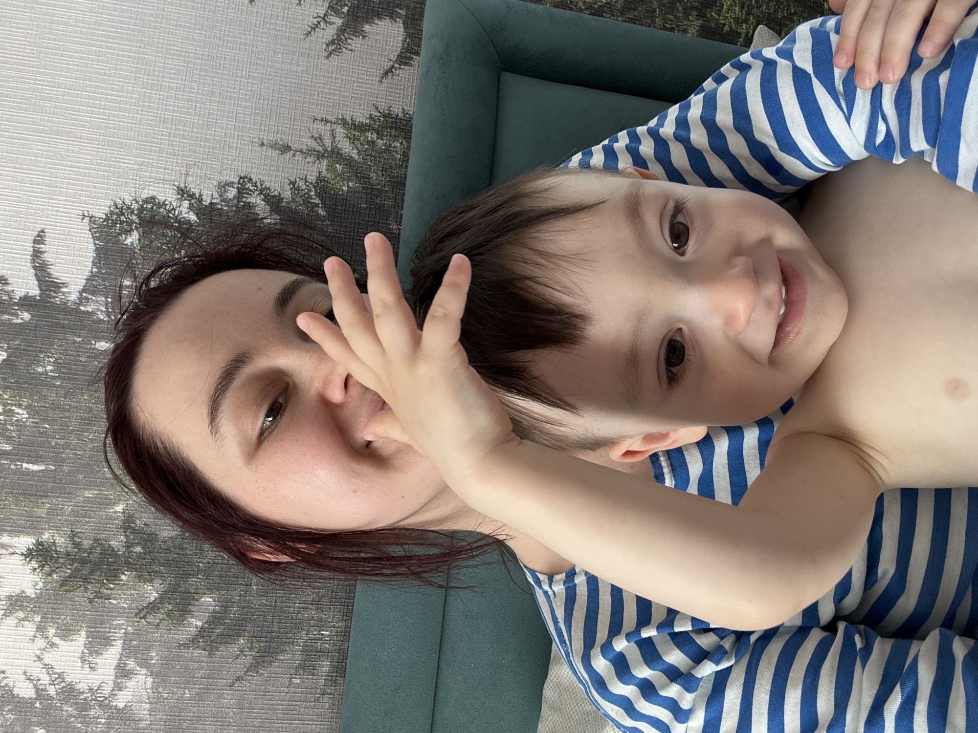 A woman and a young child are lying down. The woman has dark hair and is partially obscured by the child's hand. The child is wearing a blue and white striped shirt. The background includes a textured wall with a pattern resembling trees and a green couch. There is no information to suggest this image was taken in Izhevsk, Russia.