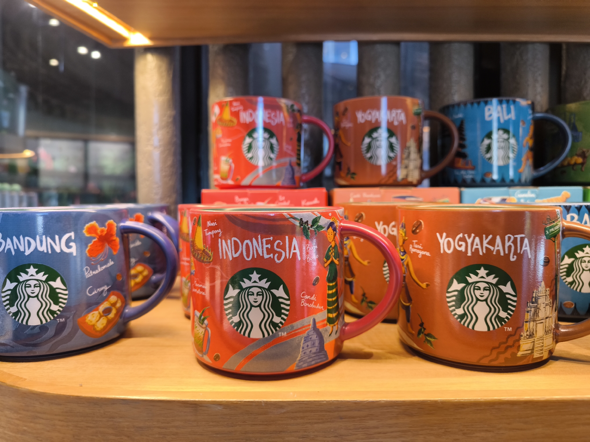 The image shows a display of Starbucks mugs, likely inside a Starbucks store. The mugs are designed to showcase different cities and regions of Indonesia, specifically "Bandung," "Indonesia," "Yogyakarta," and "Bali." Each mug features the city or region's name, along with iconic local symbols, landmarks, or cultural elements. The mugs are arranged on a light-colored wooden shelf. The mugs are new and appear to be on display for sale.