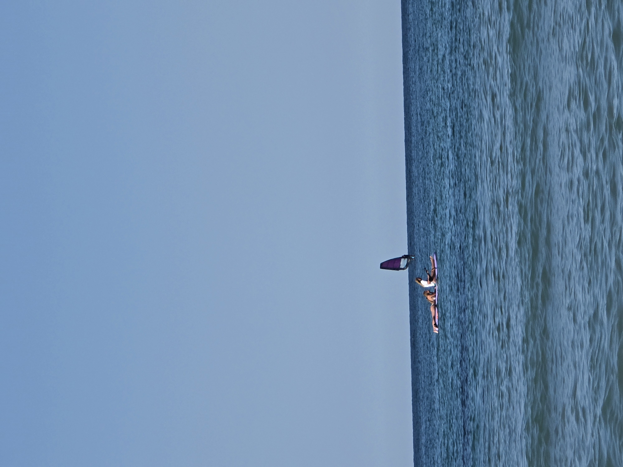 The image shows a bright, clear day at sea in Ahtopol, Bulgaria. The sky is a uniform light blue, with no clouds visible. The sea is a deep blue, with small waves creating a rippled texture. In the middle of the frame, a pink paddleboard is floating on the water. Two people are on the paddleboard. One person is lying down on their stomach, facing left, wearing a bikini. Another person is sitting up, facing right, wearing a white top and dark shorts. Attached to the back of the paddleboard is a purple sail, suggesting it is a windsurfing board that is not currently being used for windsurfing. The horizon line where the sea meets the sky is sharp and clear.