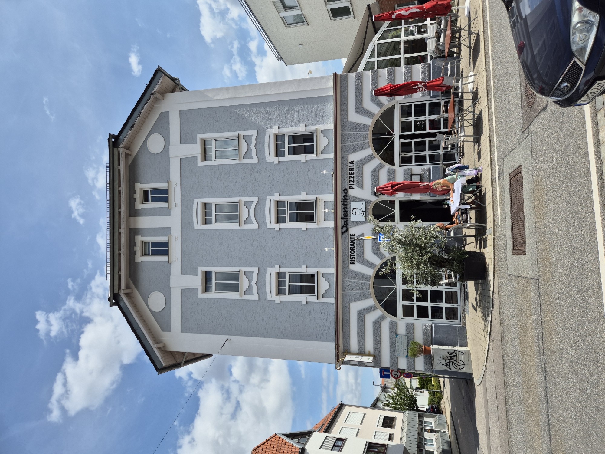 A medium shot captures a two-story building with a grey stucco facade and white trim, housing a "Valentino Ristorante Pizzeria". The building features several windows with decorative white frames on the upper floor and large arched windows with glass doors on the ground floor. Red umbrellas are set up outside the pizzeria, with tables and chairs on the sidewalk. Two women are seated at one table, one facing forward and the other looking to her right. To the left of the pizzeria, another building with a red tile roof is visible, and a street with traffic signs and a speed limit sign indicating "30" is in the foreground. A dark blue car is parked in the lower right corner of the image. The sky above is blue with scattered white clouds. The scene appears to be in Regensburg, Germany.