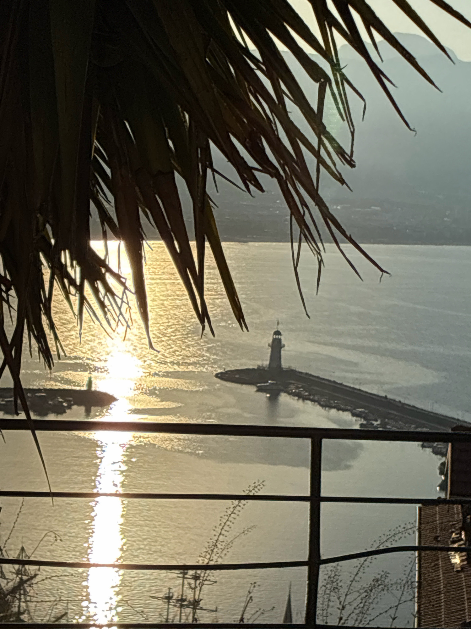A scenic view of the Alanya Marina in Alanya, Turkey, is captured, framed by the fronds of a palm tree in the foreground. The sun is low on the horizon, casting a shimmering reflection across the calm water of the sea. A lighthouse stands prominently at the end of a long pier, its white structure silhouetted against the glowing water. Several boats are docked along the pier. The background shows a hazy silhouette of mountains and the Alanya cityscape. A metal railing in the lower portion of the image suggests the vantage point is from a balcony or elevated structure. The overall atmosphere is tranquil, illuminated by the warm, golden light of the setting sun.