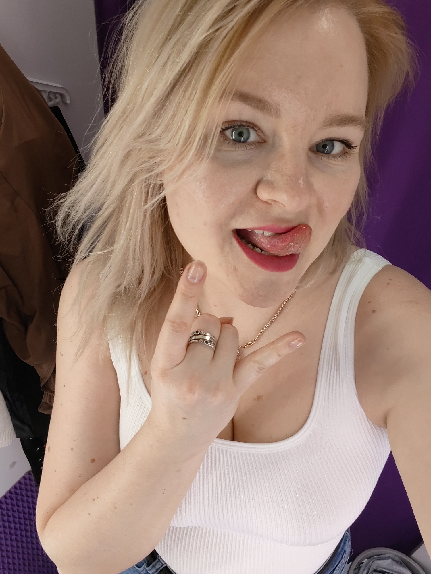 A blonde woman with blue eyes is centered in a close-up selfie. Her hair is styled in loose waves, and she wears red lipstick. She is sticking out her tongue in a playful expression, and her hand is raised, displaying a "rock on" hand gesture with a ring on her ring finger. She wears a white ribbed tank top and a gold necklace. The background includes a brown garment hanging on a rack to the left, and a purple wall to the right. No specific location in Izhevsk, Russia is discernible from the image.